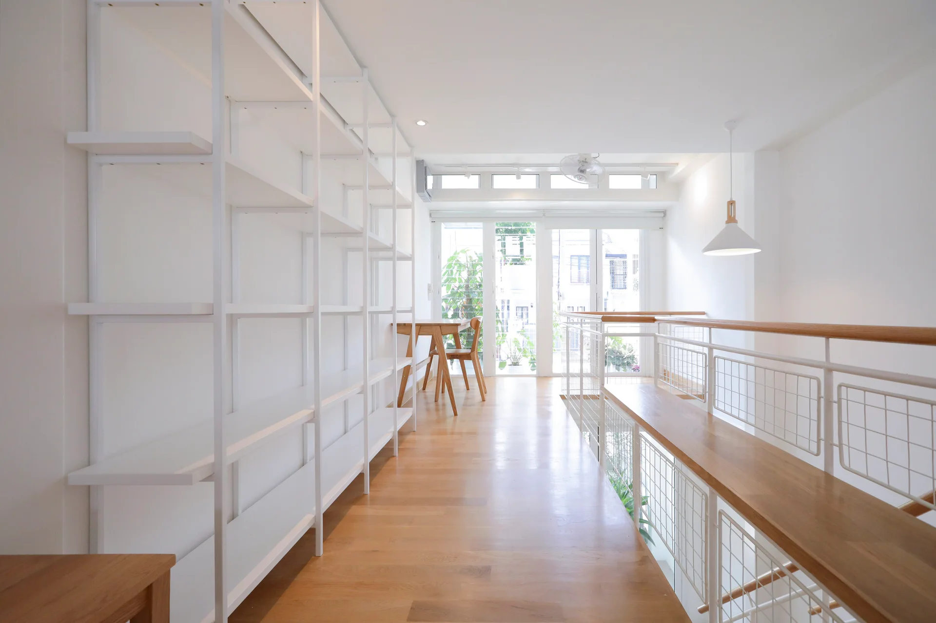 A bright residential landing featuring light wood flooring, a tall empty white metal bookshelf, and a wood-topped white metal railing.