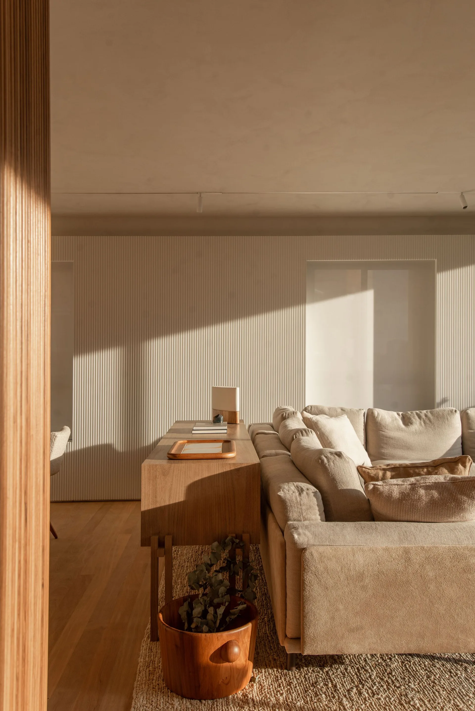 A beige fabric sofa backed by a low wooden console table sits in front of a white fluted wall with diagonal sunlight shadows.