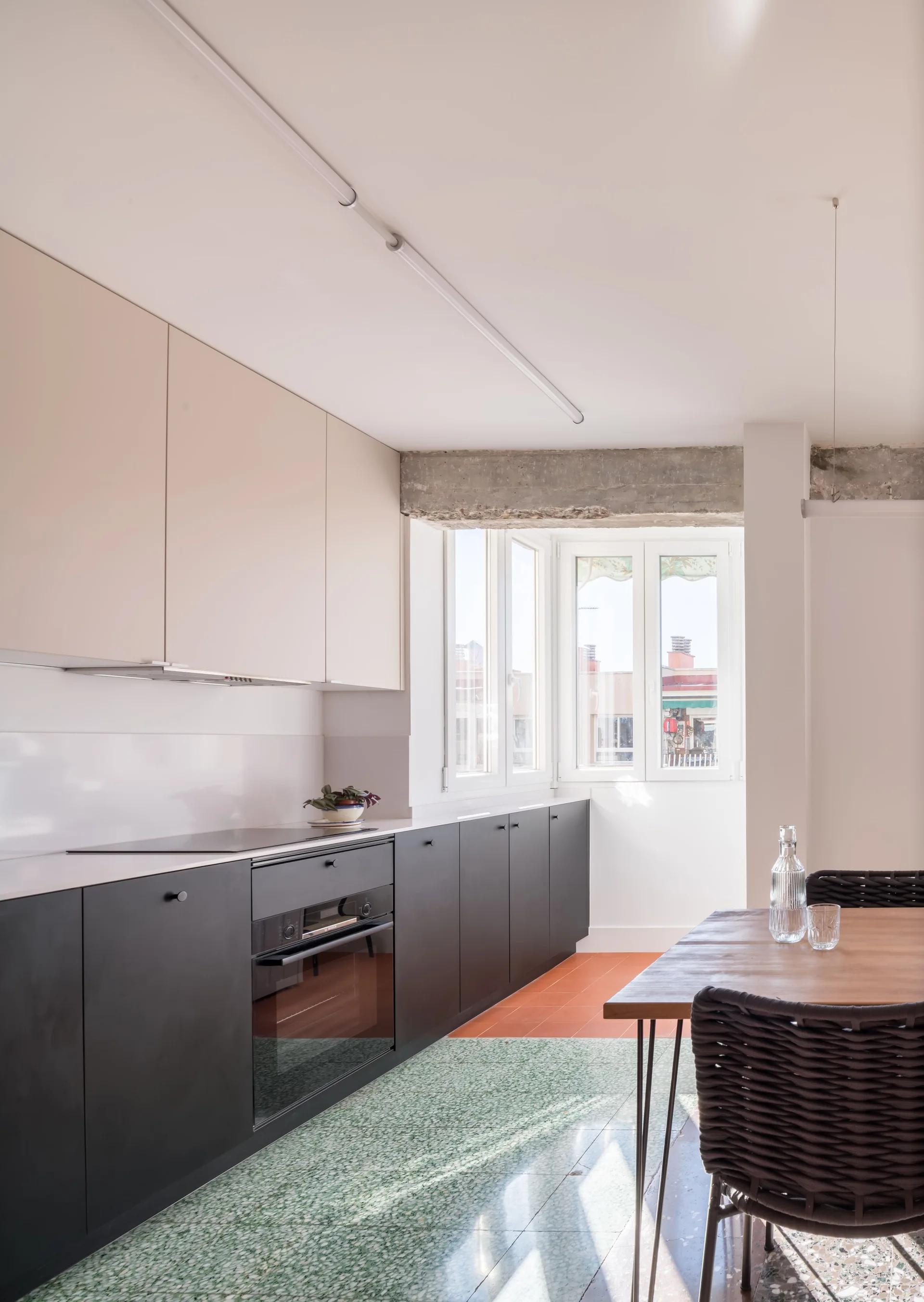 A modern kitchen with black and cream cabinets features green terrazzo flooring, a wooden dining table, and an exposed concrete beam above the window.