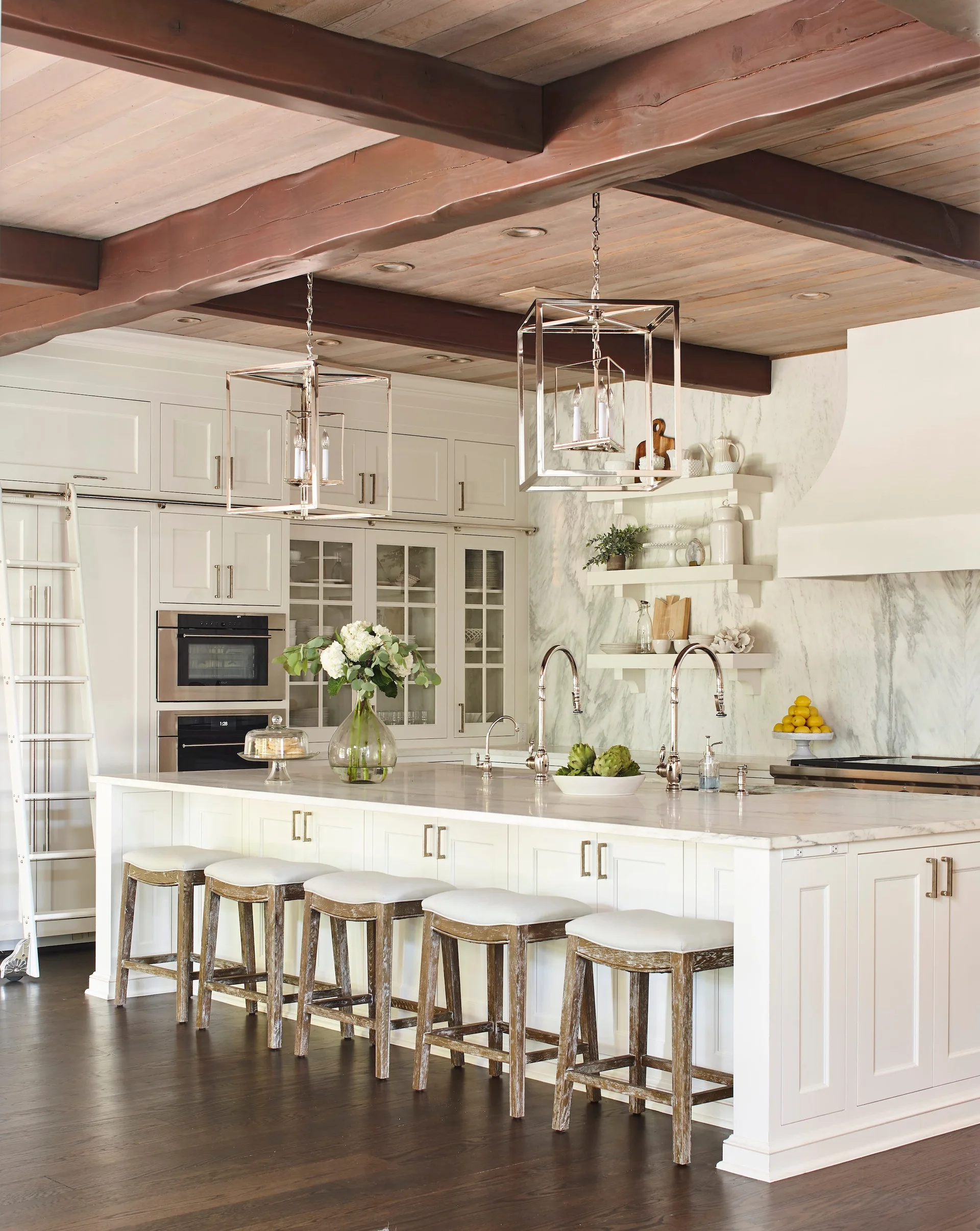 A bright kitchen features a large white island with barstools beneath a wood plank ceiling with dark exposed beams and two lantern pendant lights.