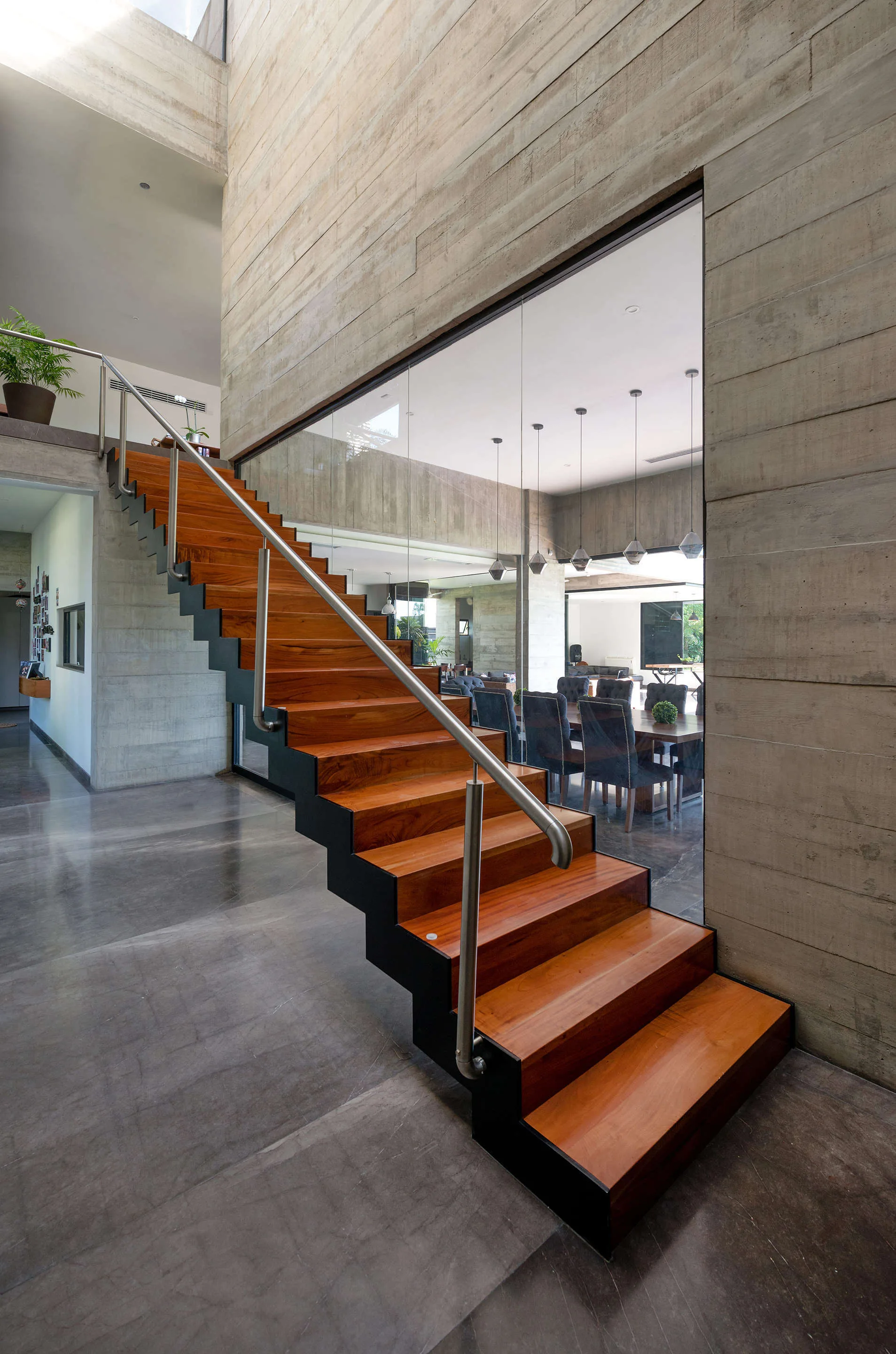 A modern wooden staircase with a metal railing stands next to a board-formed concrete wall and a glass partition looking into a dining area.