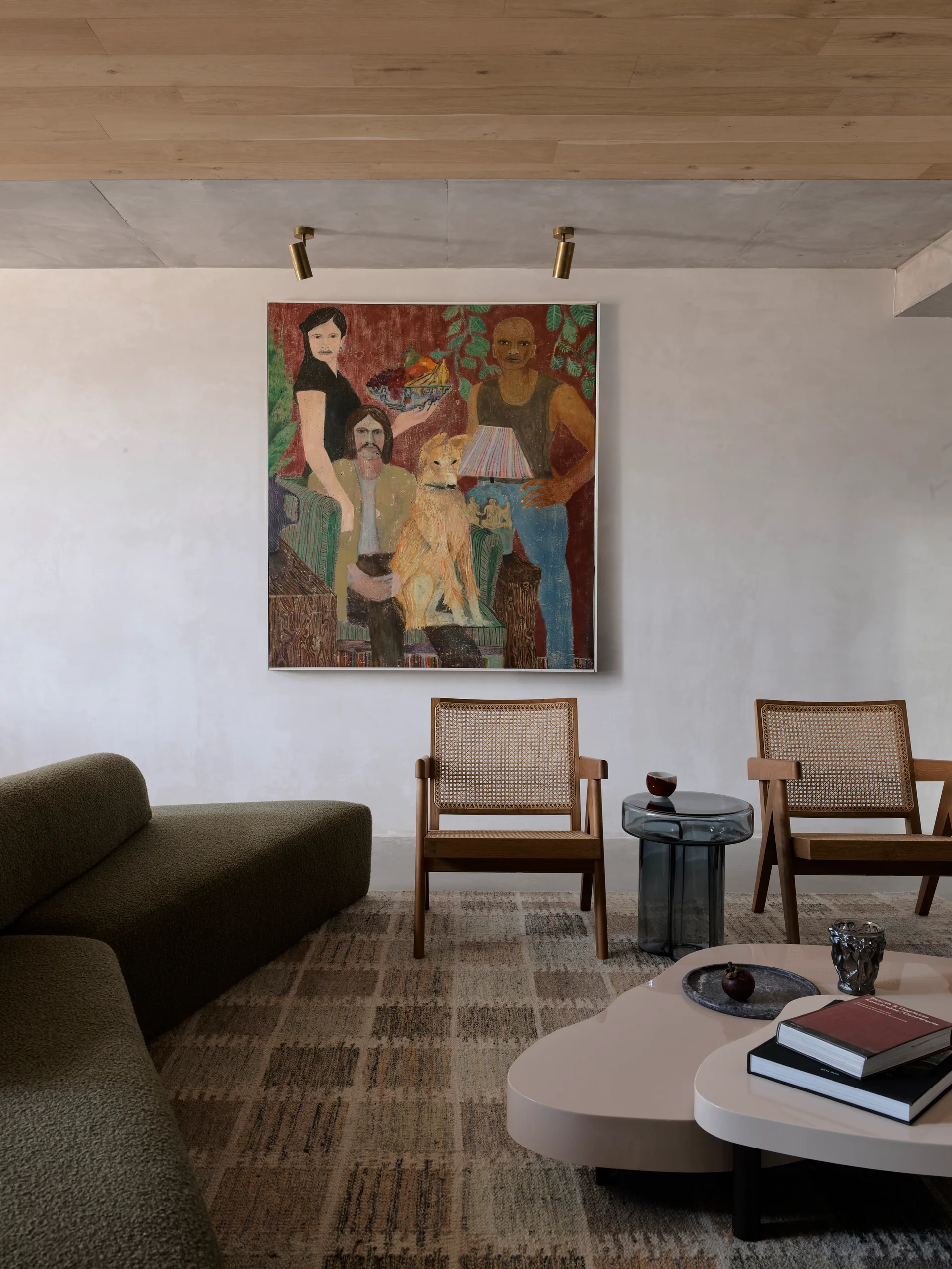 A living room with an olive green sofa, two wooden cane armchairs, a low organic coffee table, and a large painting on a plaster wall.