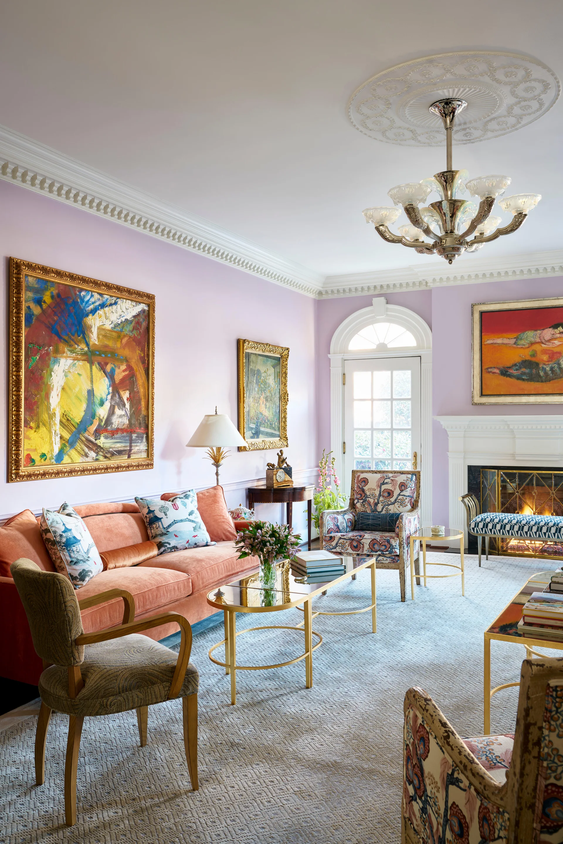A formal living room features a coral velvet sofa, light lavender walls, patterned armchairs, and a fireplace framed by gilded artwork.