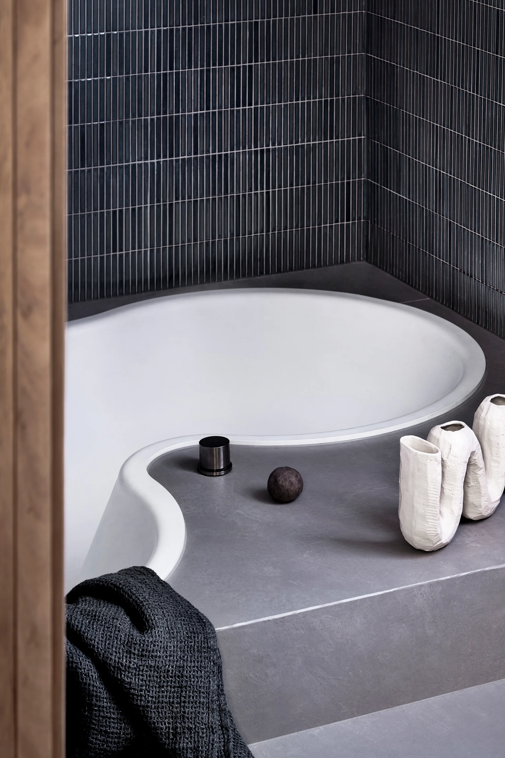 A curved white built-in bathtub is surrounded by a matte grey concrete deck and dark blue vertical mosaic wall tiles.