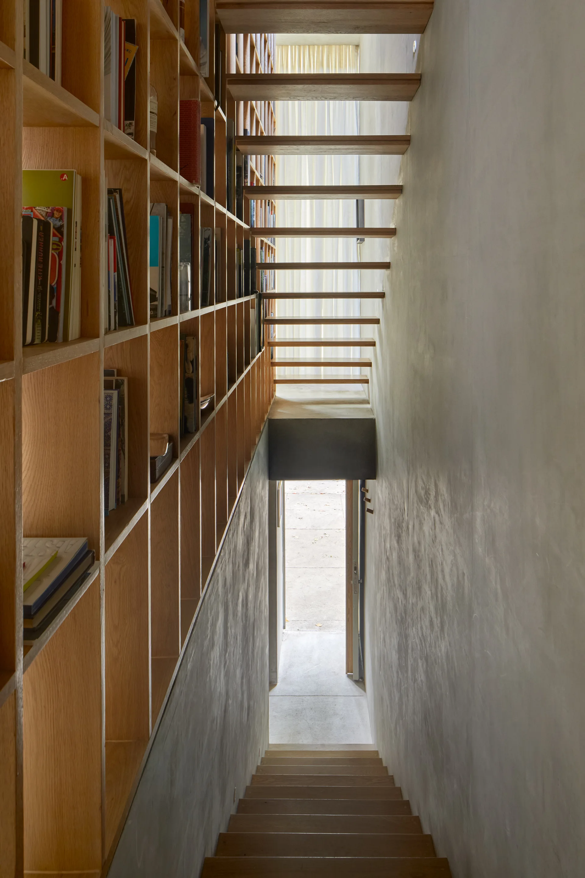 A narrow stairwell features floating wood steps, a floor-to-ceiling wooden grid bookcase filled with books, and a smooth gray plaster wall.