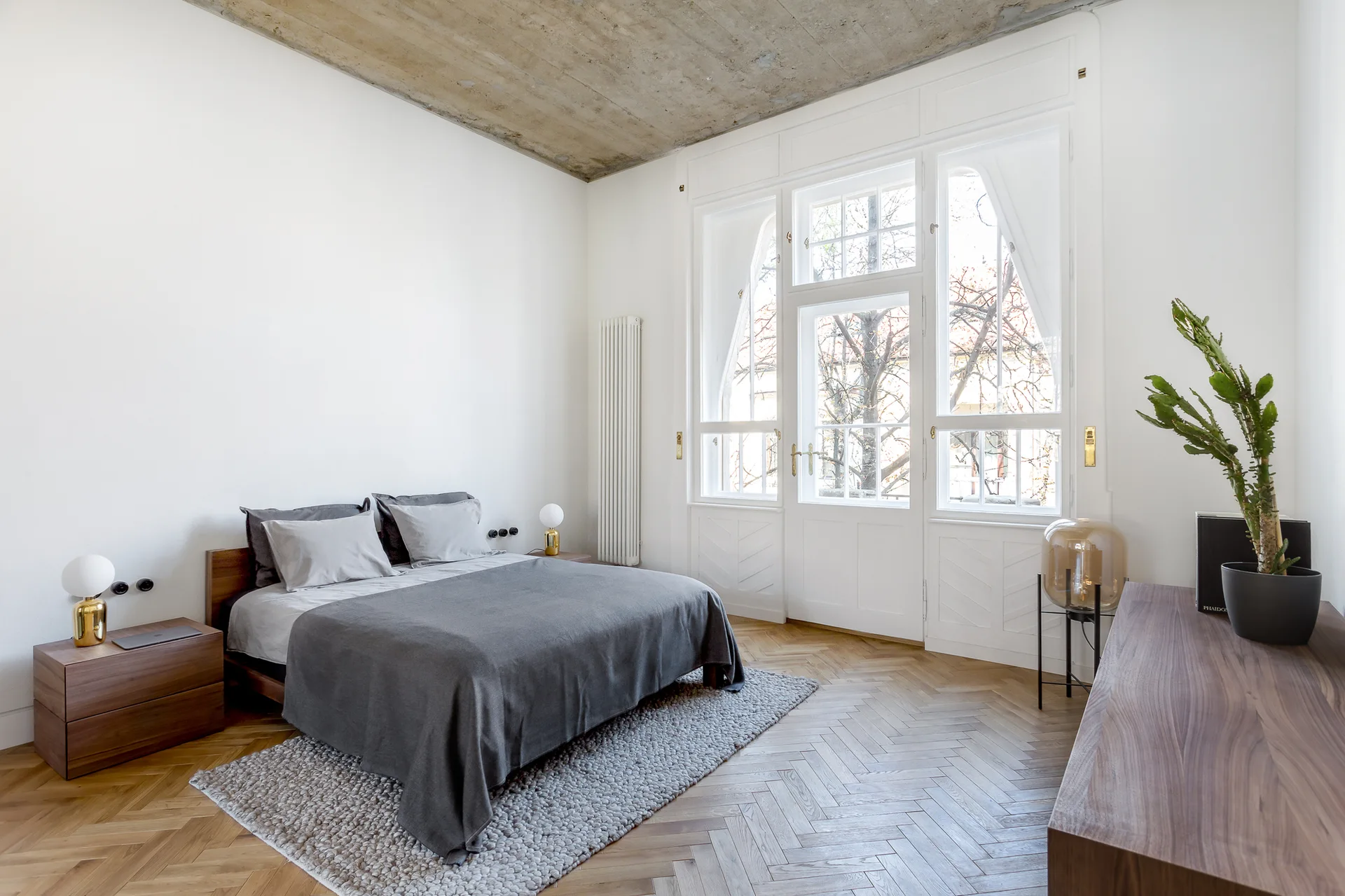 A minimalist bedroom featuring a raw concrete ceiling, a grey bed on a herringbone parquet floor, and a tall white window.