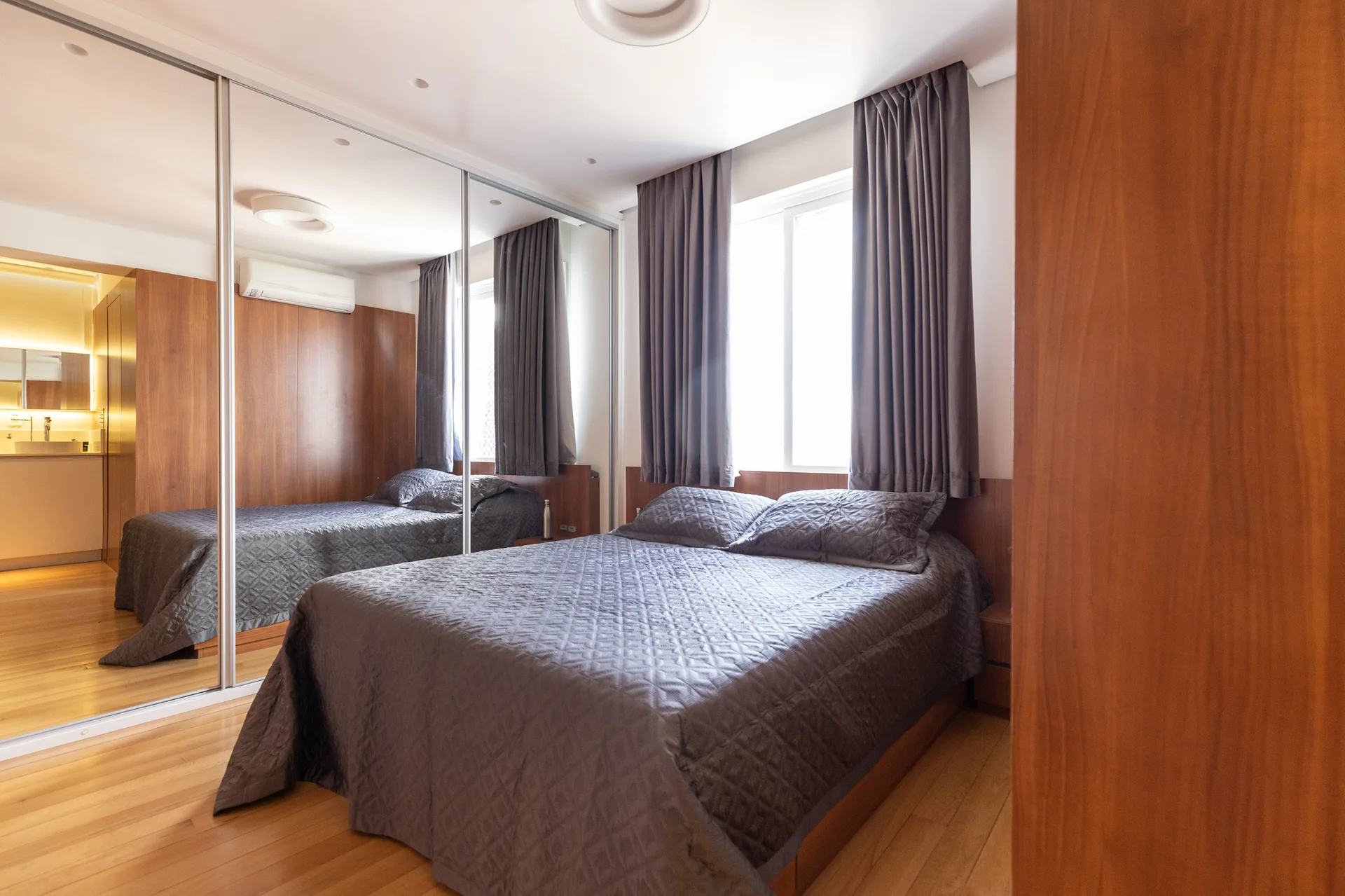 A compact bedroom features a grey quilted bed, warm wood paneling, and a large mirrored wardrobe reflecting the room.