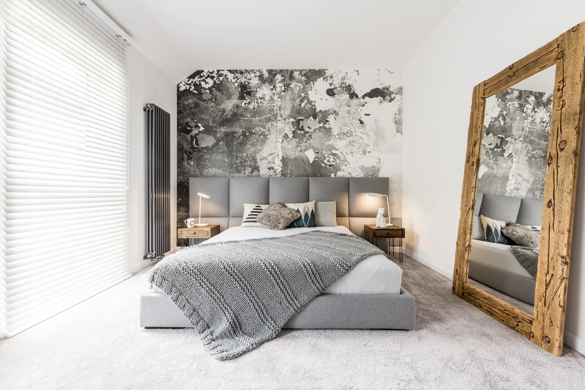 A grey upholstered bed sits against a black and white distressed mural next to a large rustic wood-framed floor mirror in a modern bedroom.