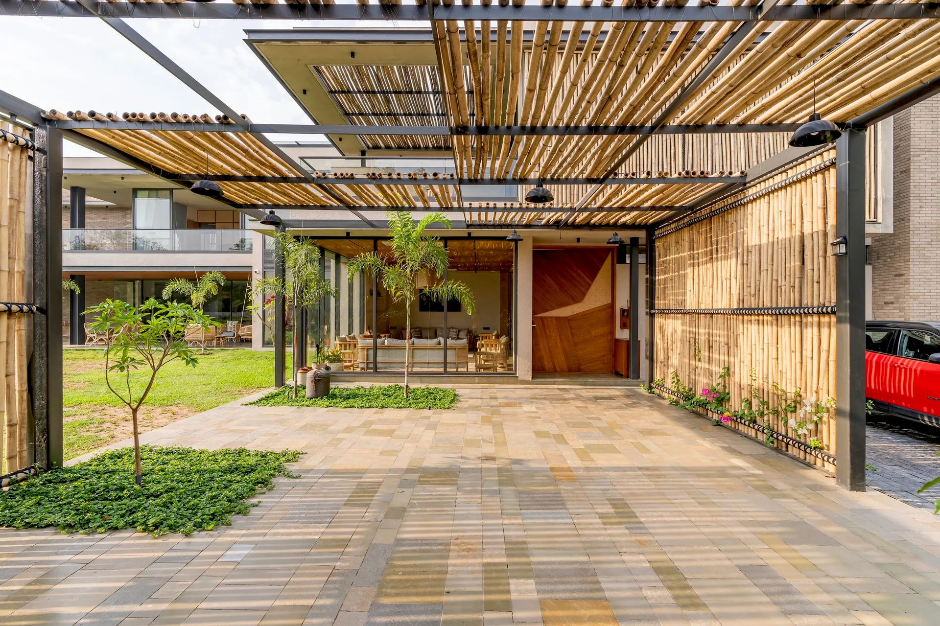 A paved stone patio features a large bamboo pergola on a black metal frame, integrated planters with small trees, and a vertical bamboo screen.