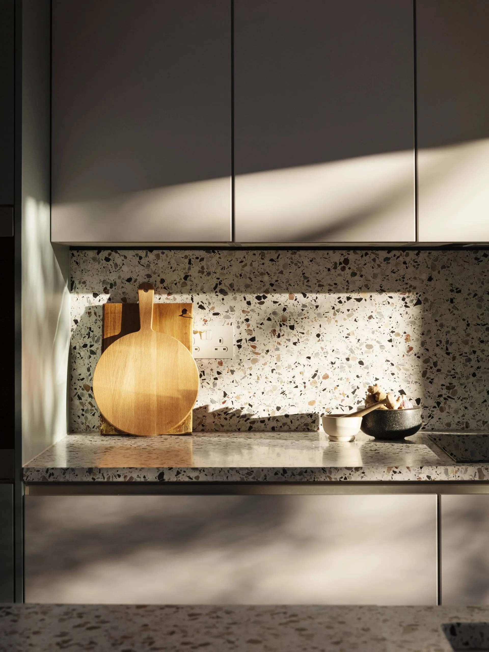 A close-up of a kitchen with light grey handleless cabinets, a speckled terrazzo backsplash and countertop, and wooden cutting boards under direct sunlight.