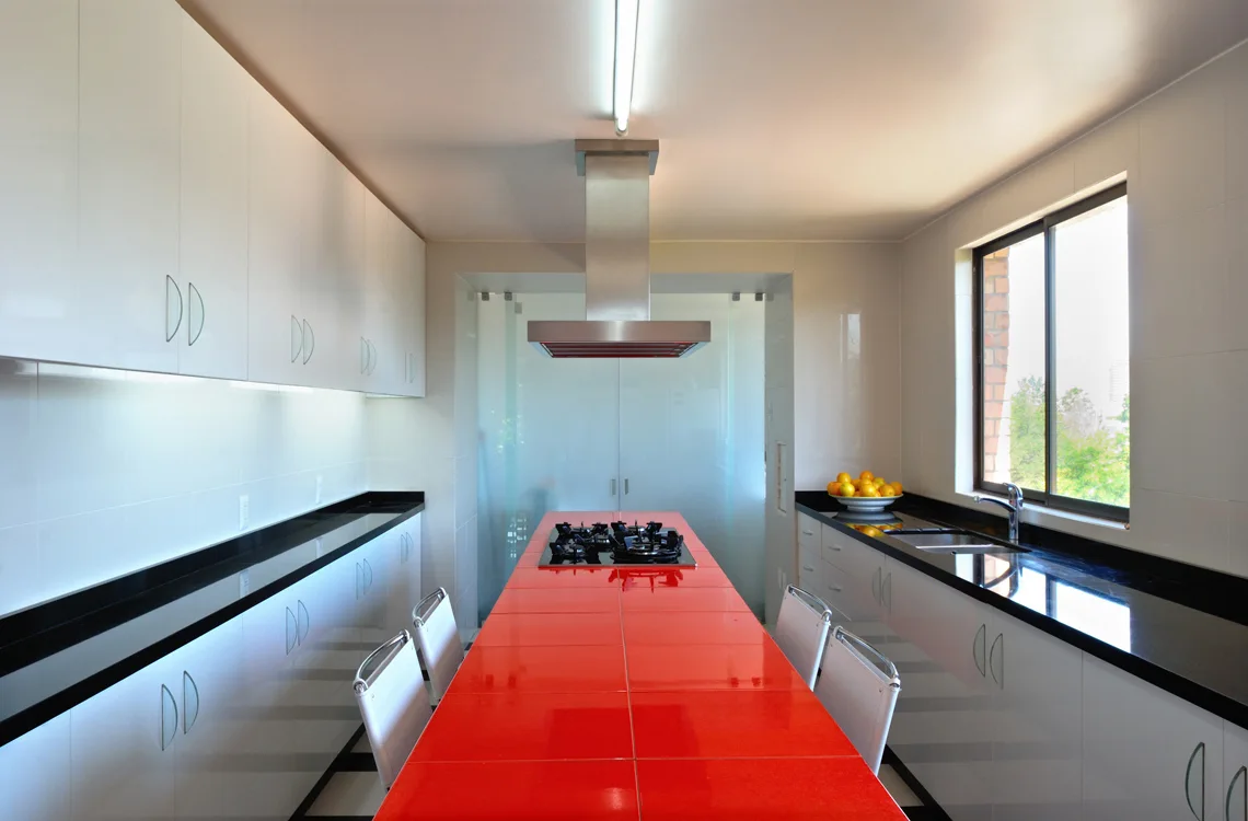 A symmetrical galley kitchen featuring glossy white cabinets, black countertops, and a long kitchen island clad in vibrant red ceramic tiles.