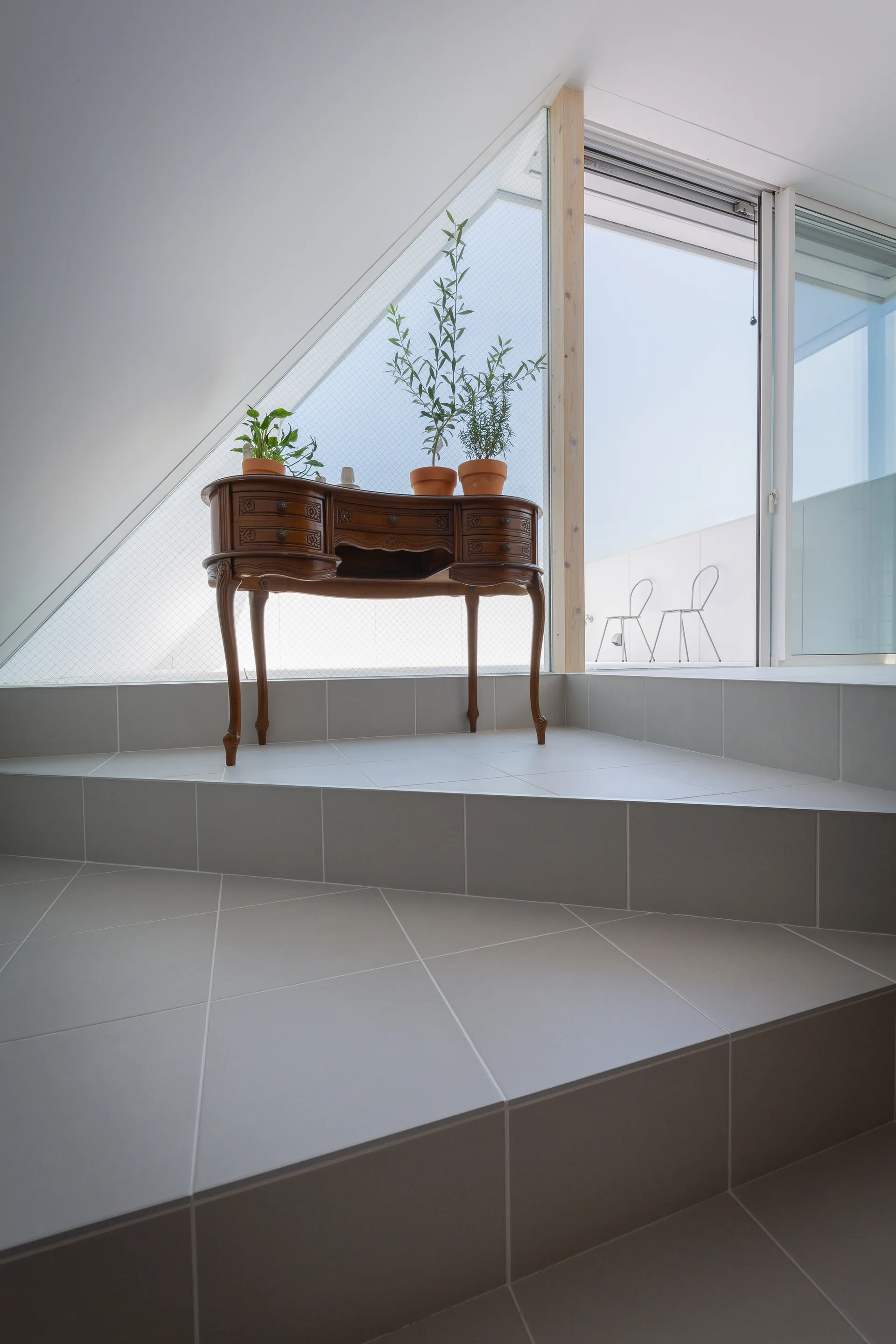 An antique wooden vanity with potted plants sits on a gray tiled staircase landing beside a large slanted window looking out to a balcony.
