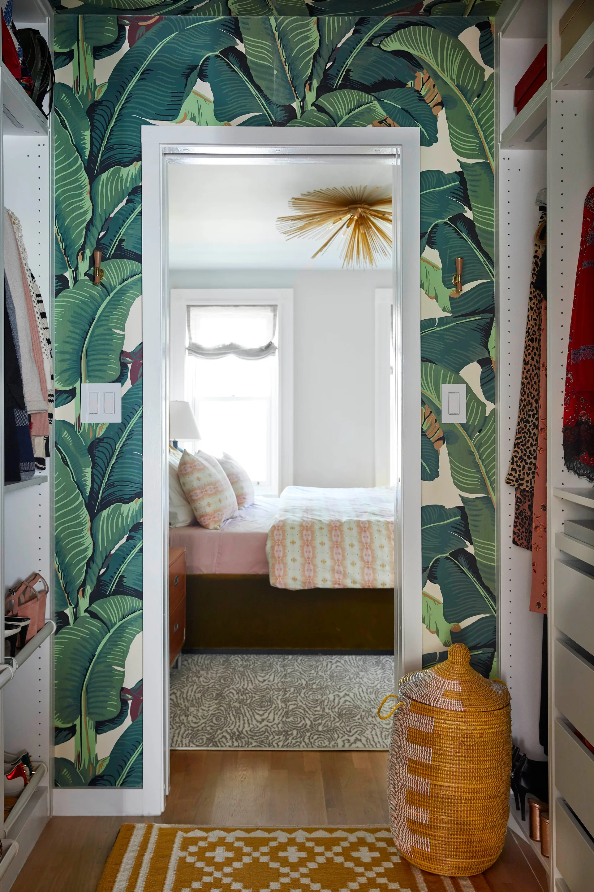 A view from a walk-in closet with green banana leaf wallpaper looking into a bedroom with a bed and a gold sunburst chandelier.