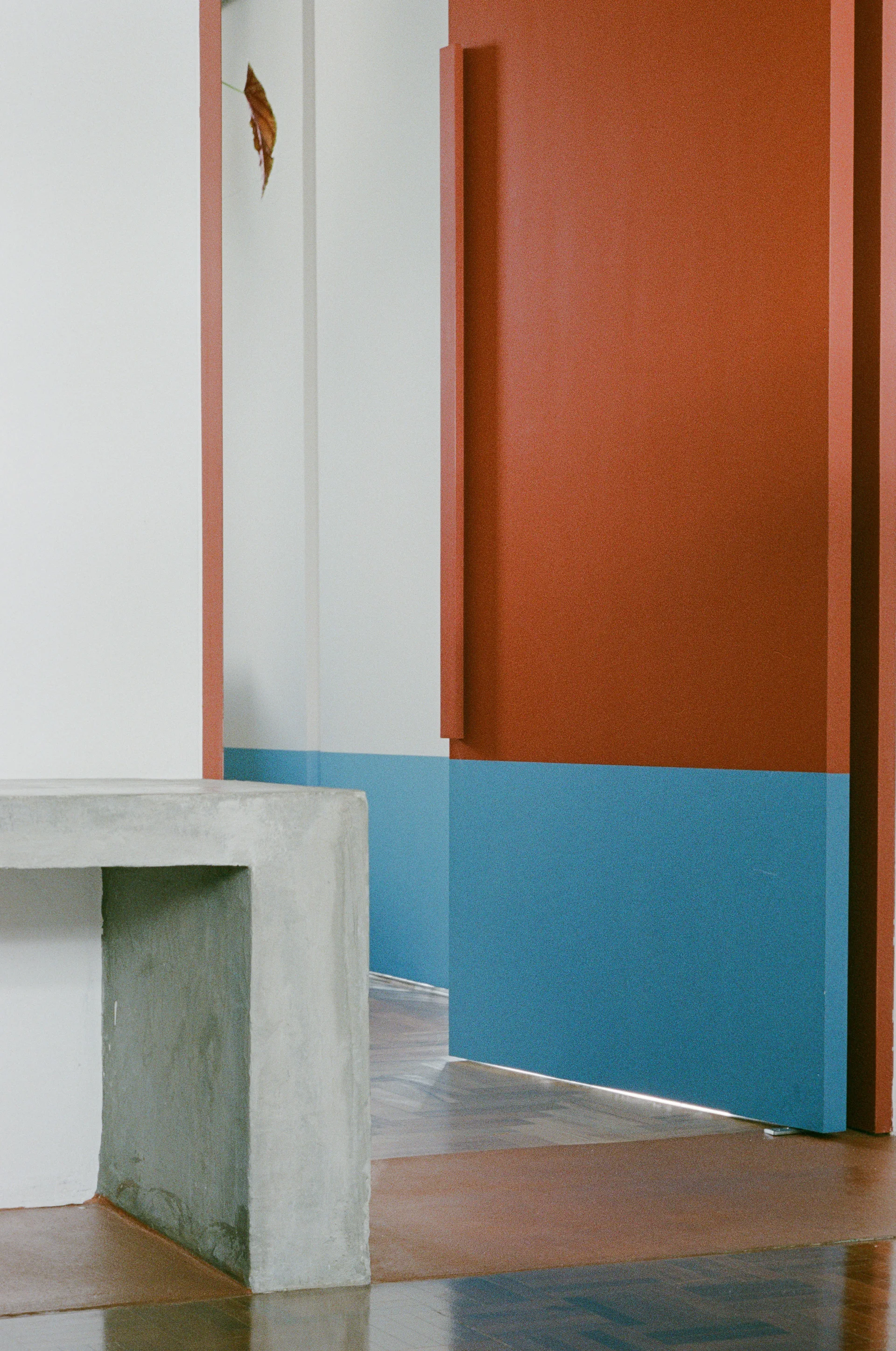 A minimalist hallway featuring a grey concrete console table in the foreground beside a modern terracotta door painted with a blue base band.