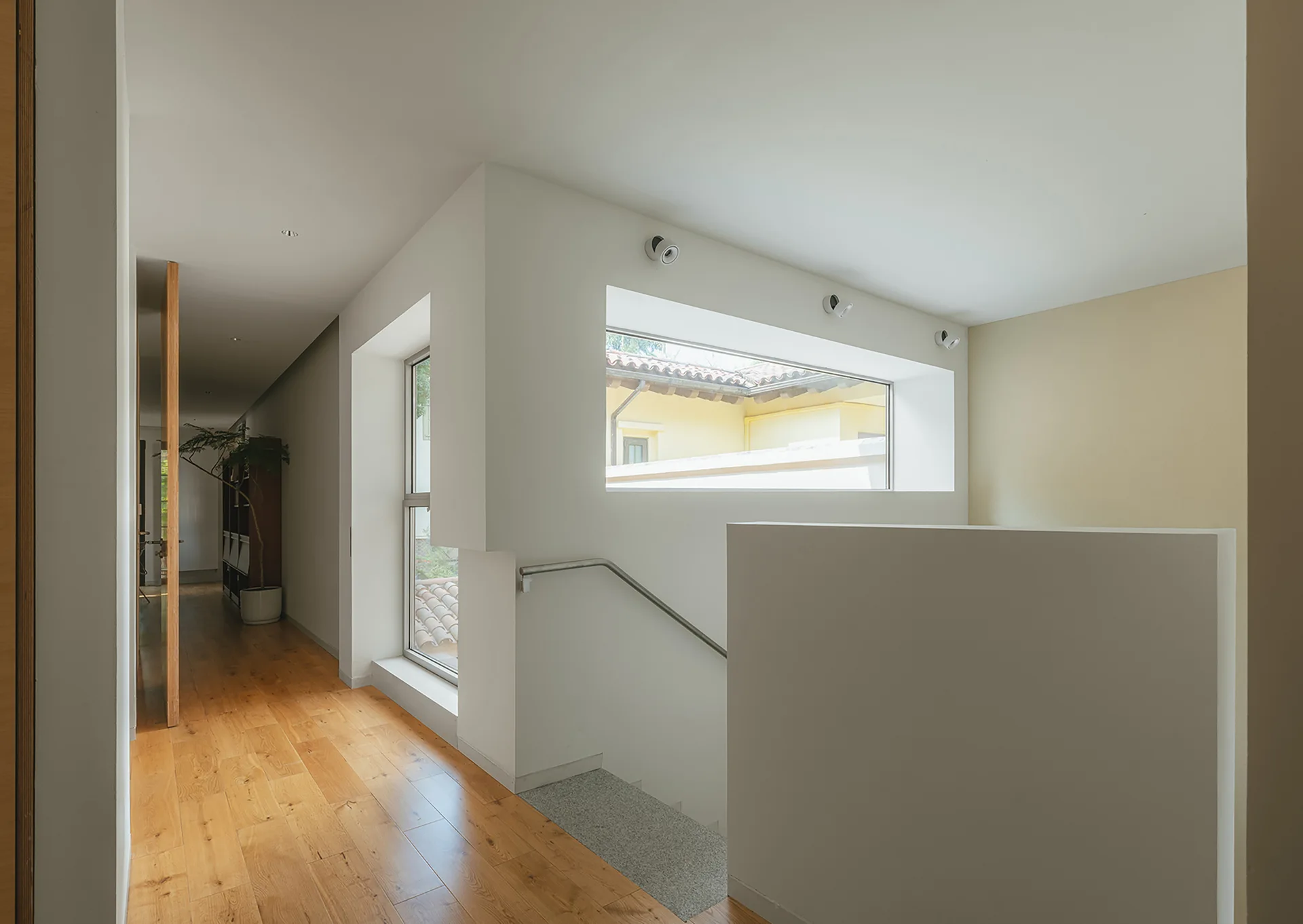 A minimalist hallway and stairwell landing featuring clean white walls, light wood flooring, and deeply recessed rectangular windows.
