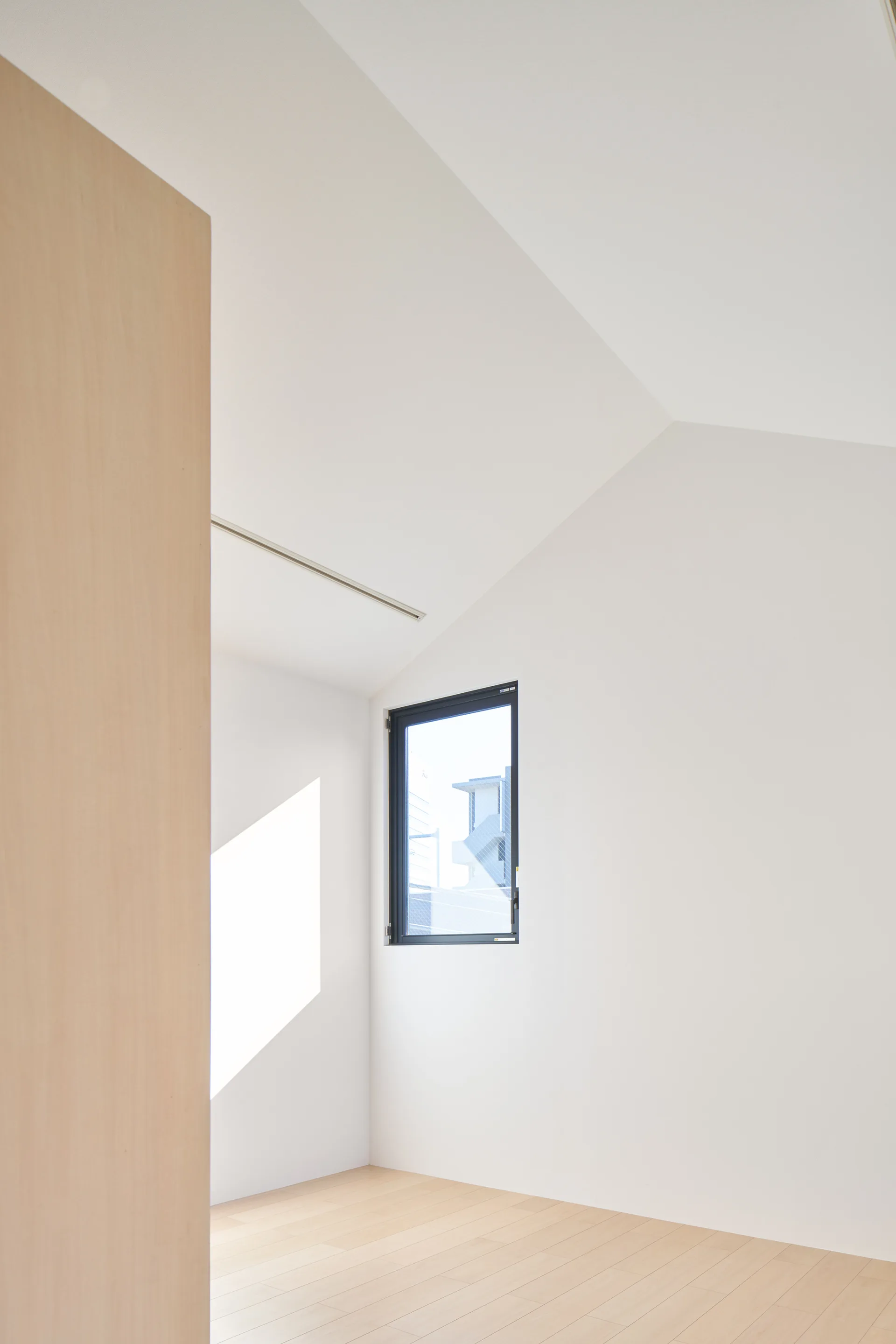A minimalist empty room features bright white walls, a sloped ceiling, light wood plank flooring, and a single black-framed square window.