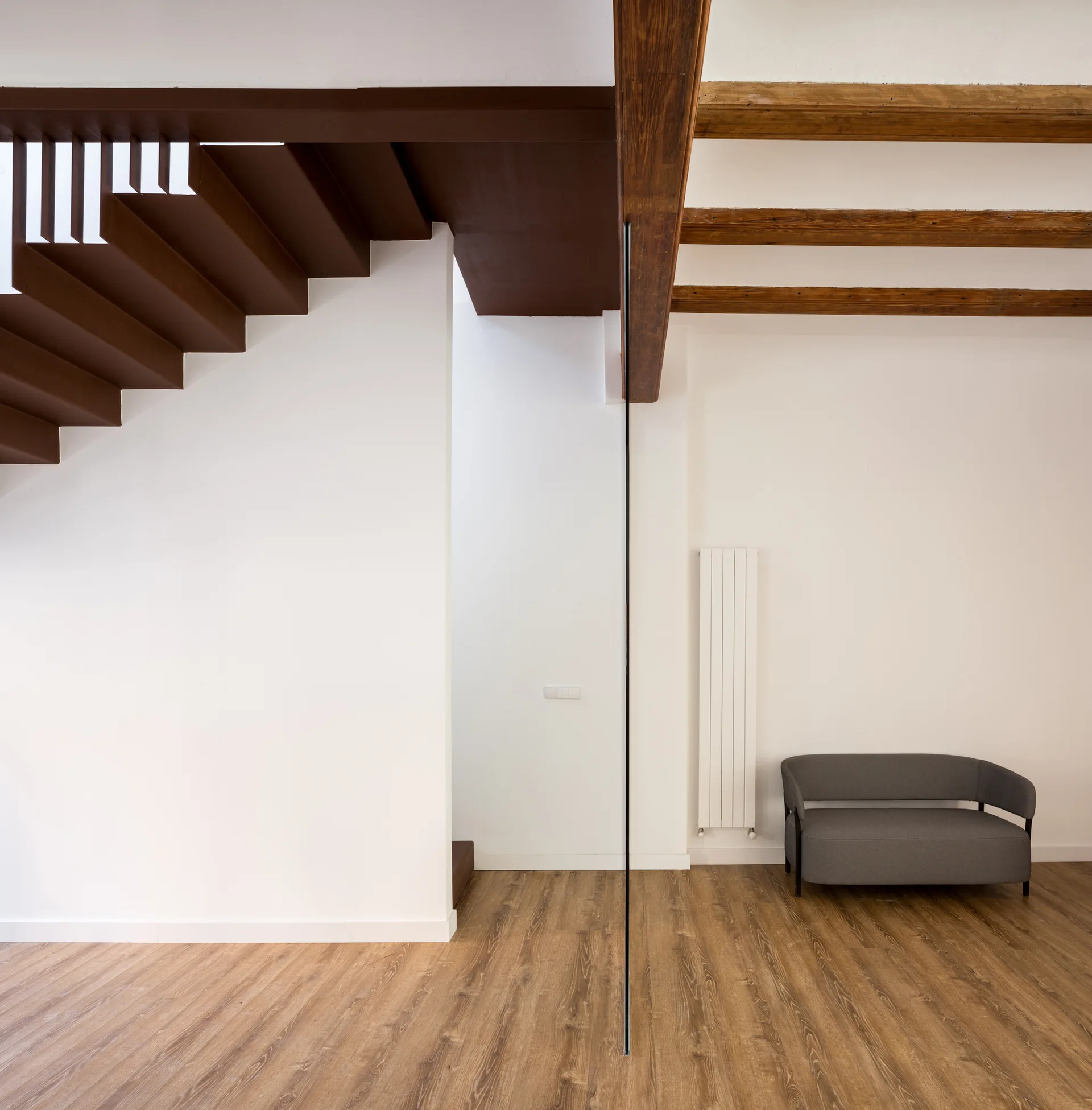 A minimalist entryway features a dark brown staircase, a tall vertical glass partition, exposed wooden ceiling beams, and a grey loveseat.