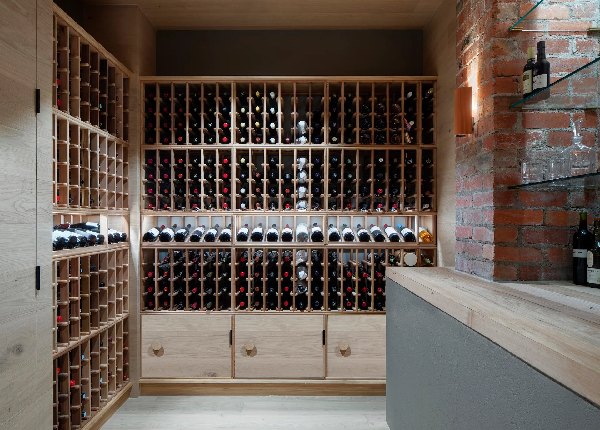 Custom light wood wine racking holds rows of bottles alongside an exposed brick wall with floating glass shelves and a warm wall sconce.