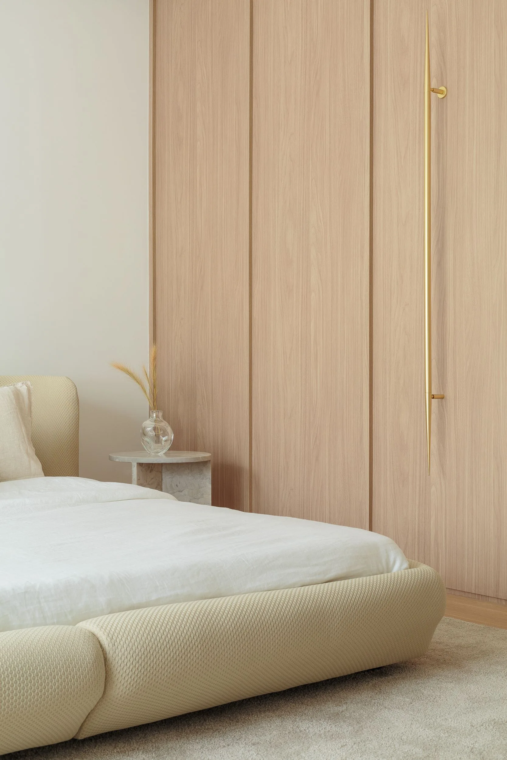A minimalist bedroom features an upholstered beige bed, a small stone side table, and built-in light wood wardrobe doors with a long gold handle.