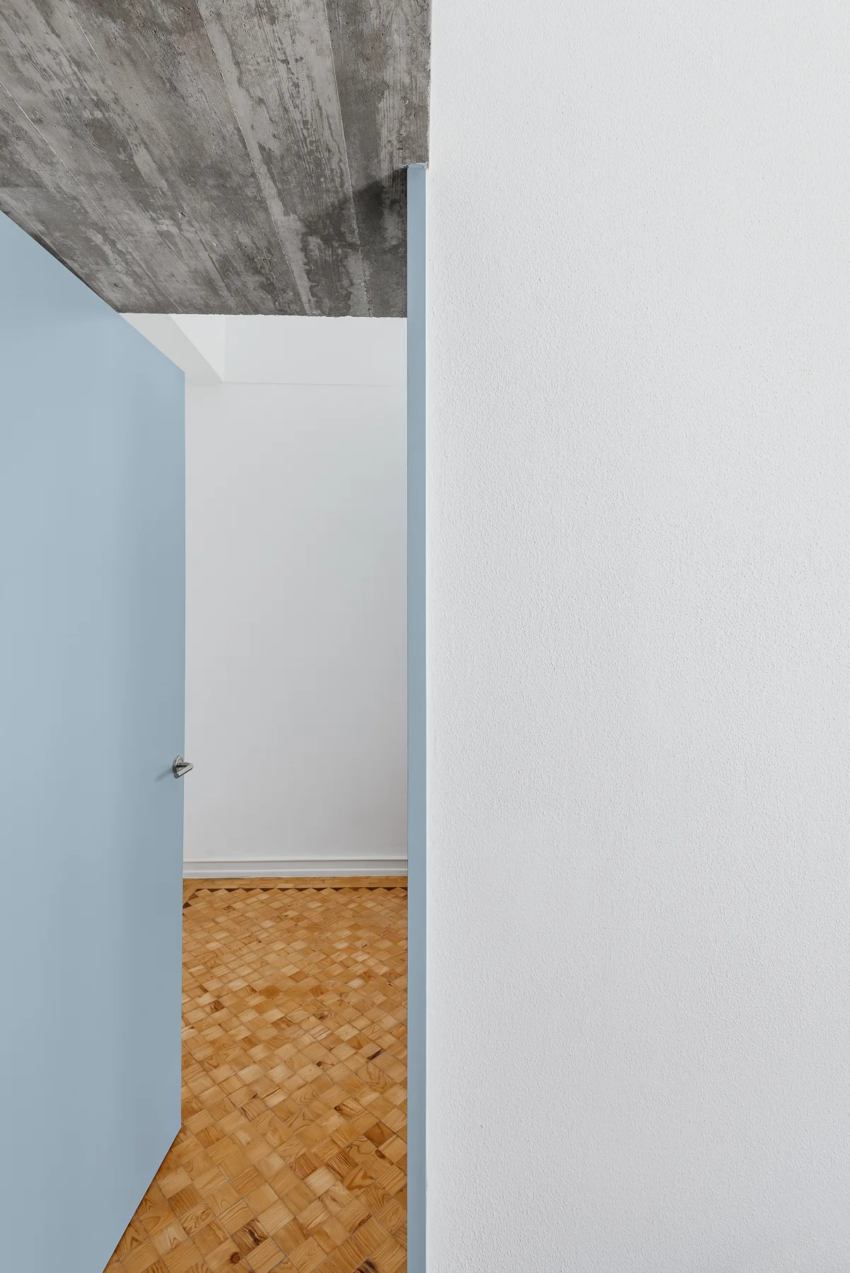 An open light blue door leads into a white room with geometric wood parquet flooring and an exposed raw concrete ceiling.