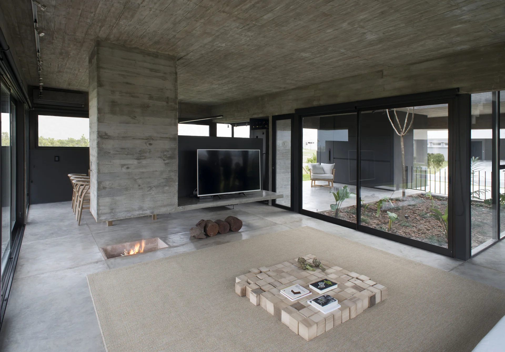 A minimalist living room features exposed concrete walls, a sunken fireplace in the floor, and a low wooden coffee table near sliding glass doors.