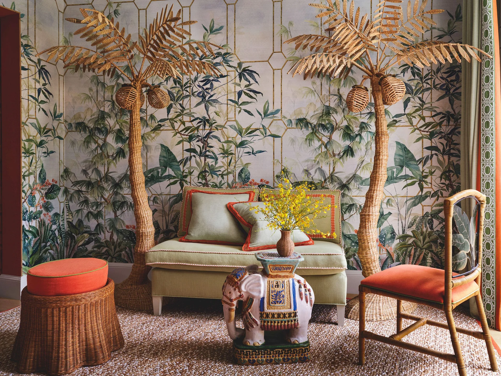 A sage green sofa sits against botanical trellis wallpaper, flanked by two woven rattan palm tree sculptures and an elephant side table on a textured rug.