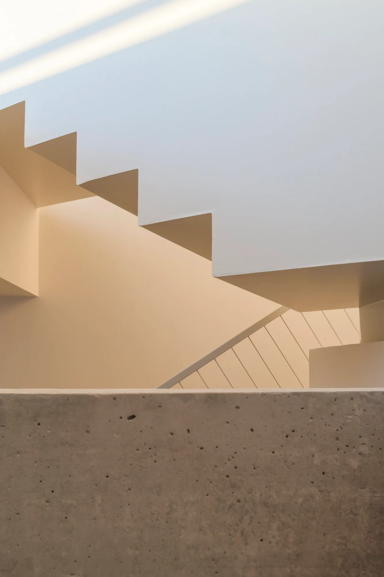 A sharp geometric white staircase rises above a textured grey raw concrete wall in a minimalist modern interior.