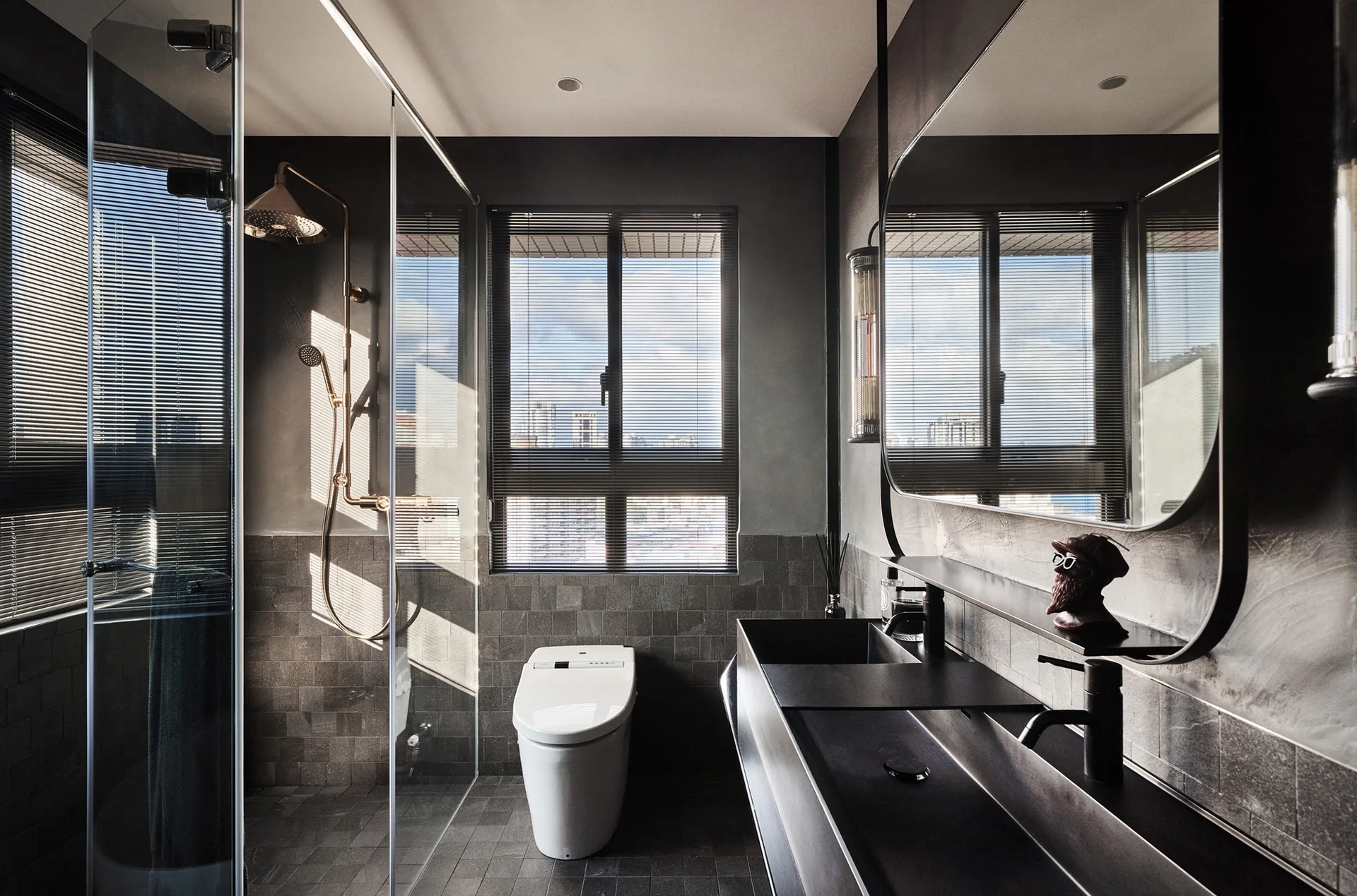A modern dark grey bathroom features a matte black trough sink, a white smart toilet, and a glass-enclosed shower with copper fixtures.