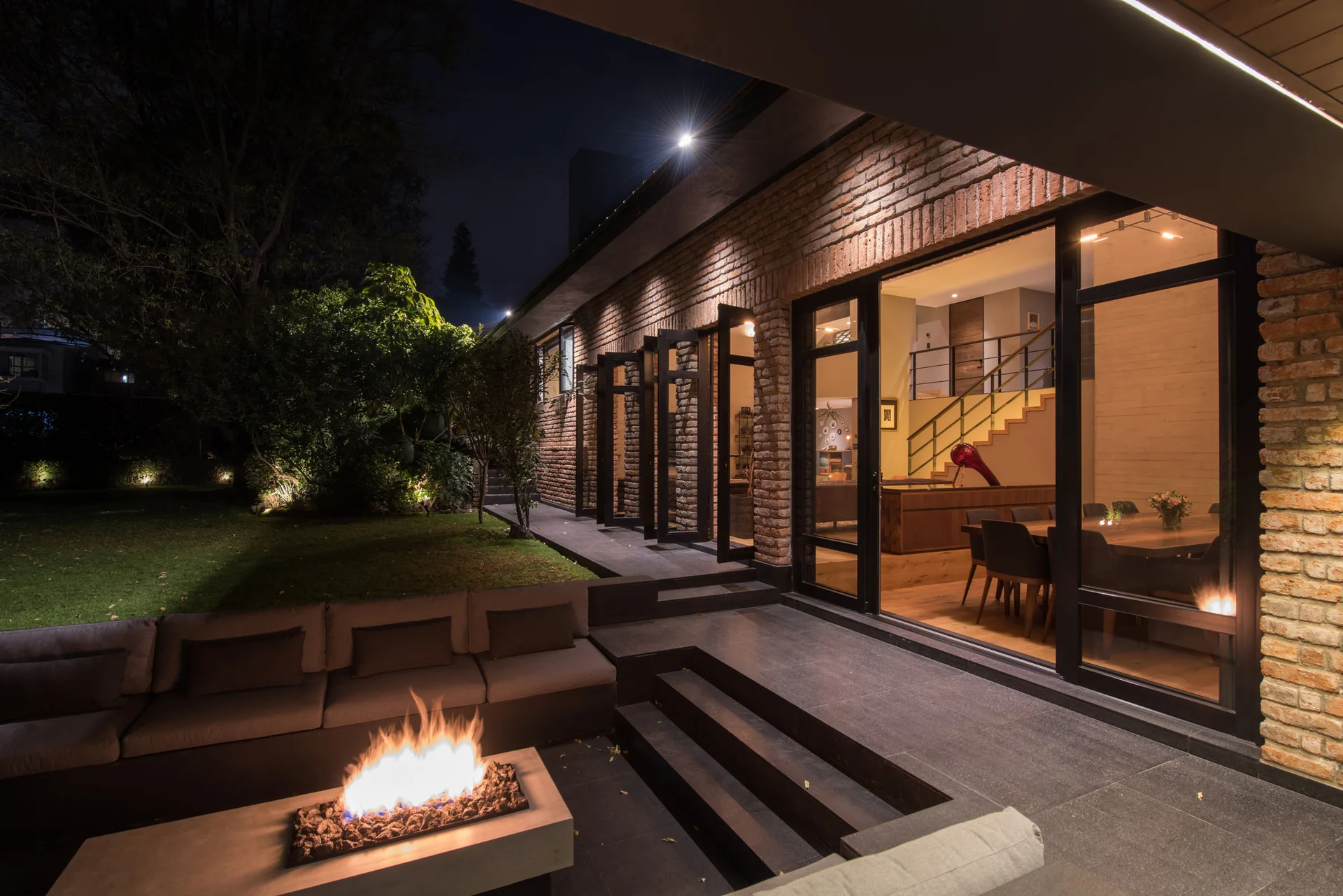 A modern brick patio at night features a sunken concrete fire pit, built-in sectional sofa, and large glass doors looking into a lit dining room.