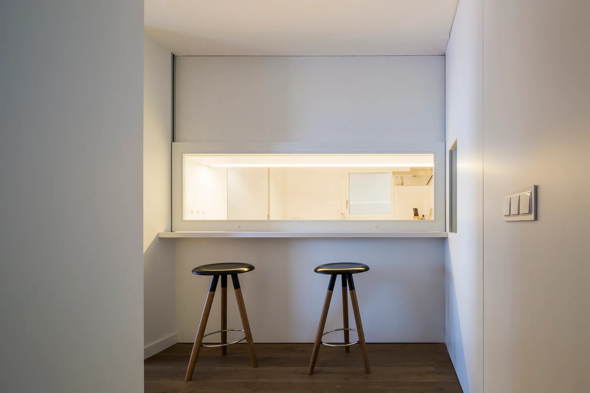 Two minimalist bar stools with tapered wooden legs stand before a white kitchen pass-through window over dark wood floors.
