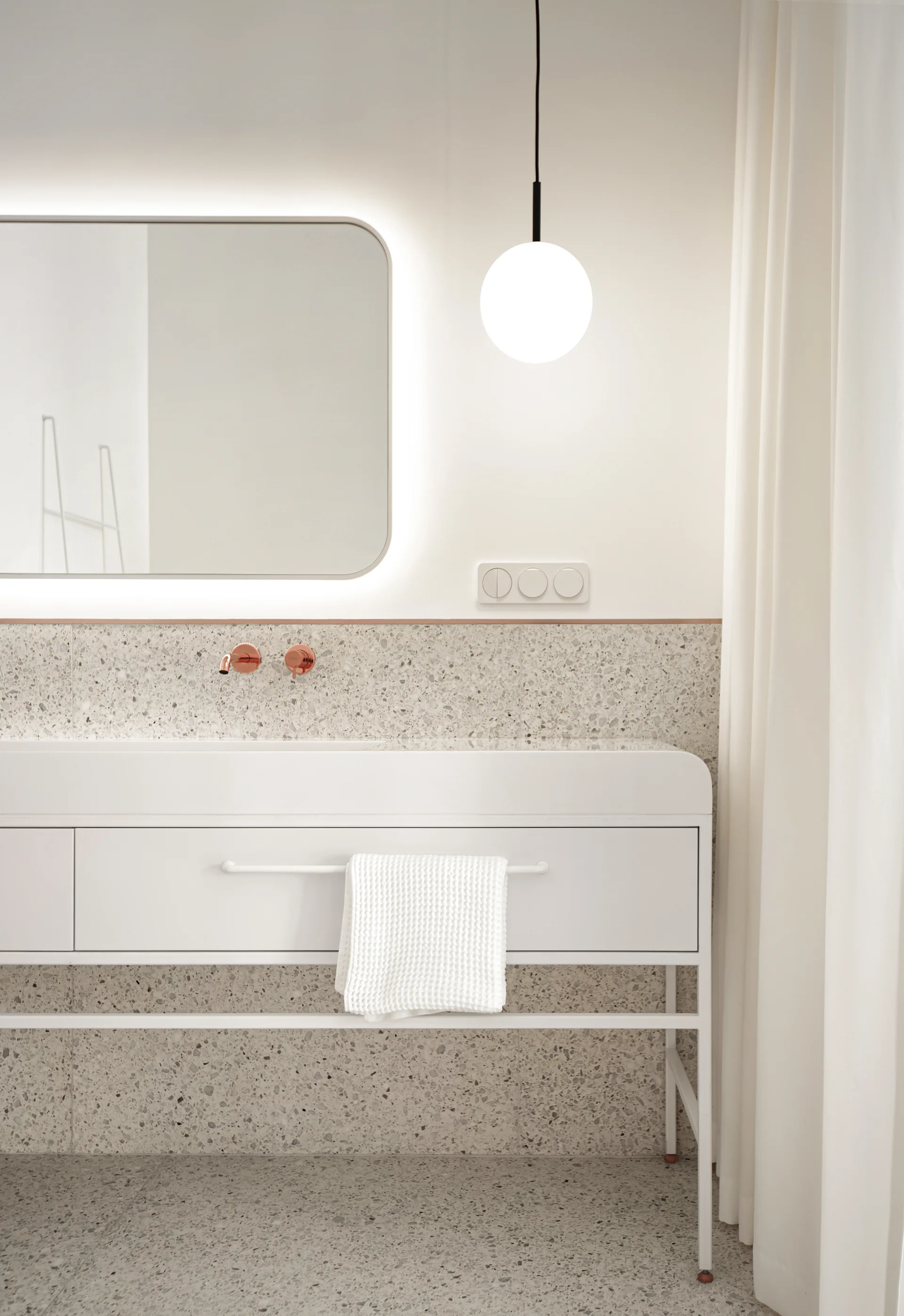 A minimalist white bathroom vanity features a speckled terrazzo backsplash and floor, paired with a backlit mirror and copper-finished faucet.