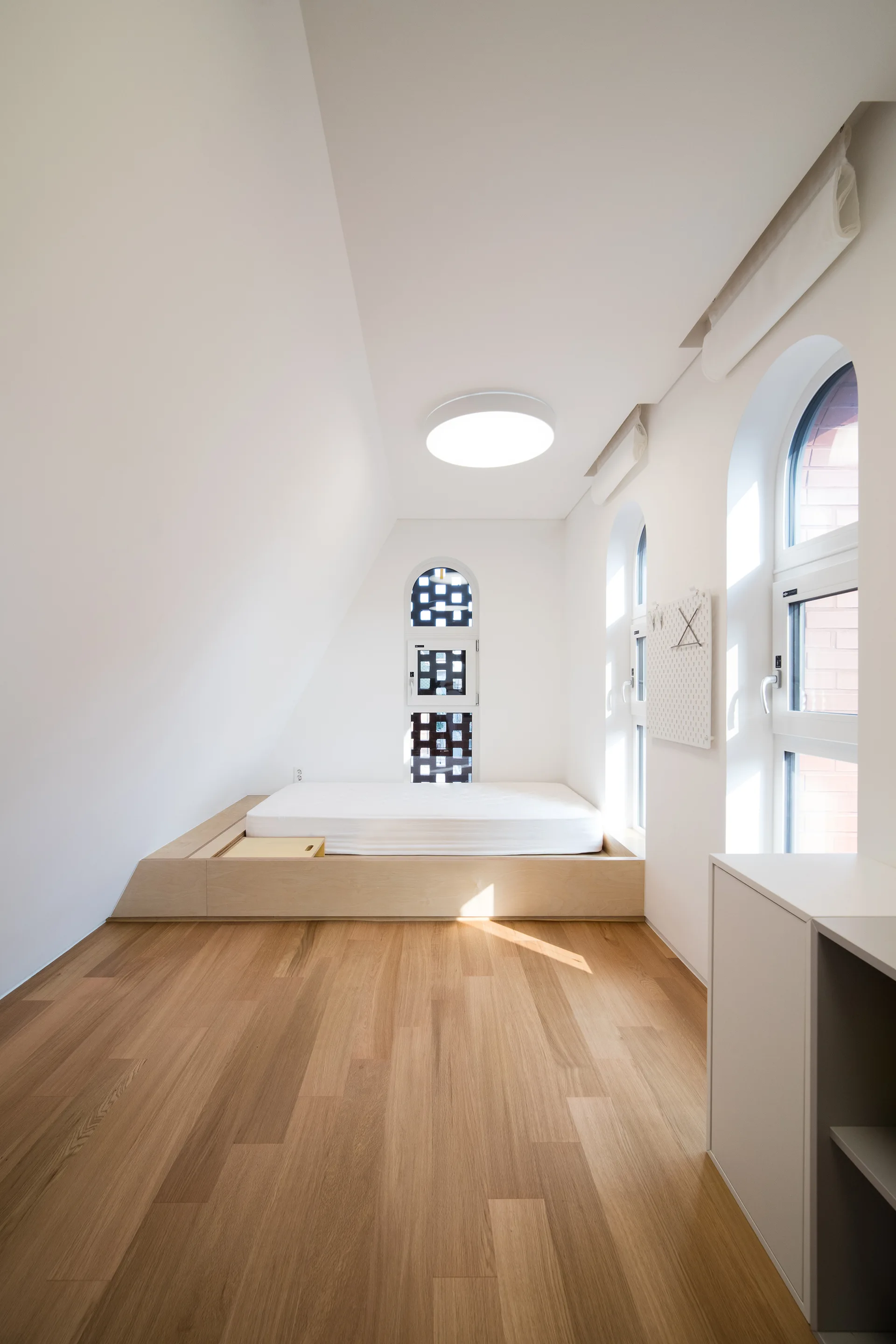 A minimalist attic bedroom with a light wood floor, white walls, and a low custom wooden bed platform positioned below an arched window.