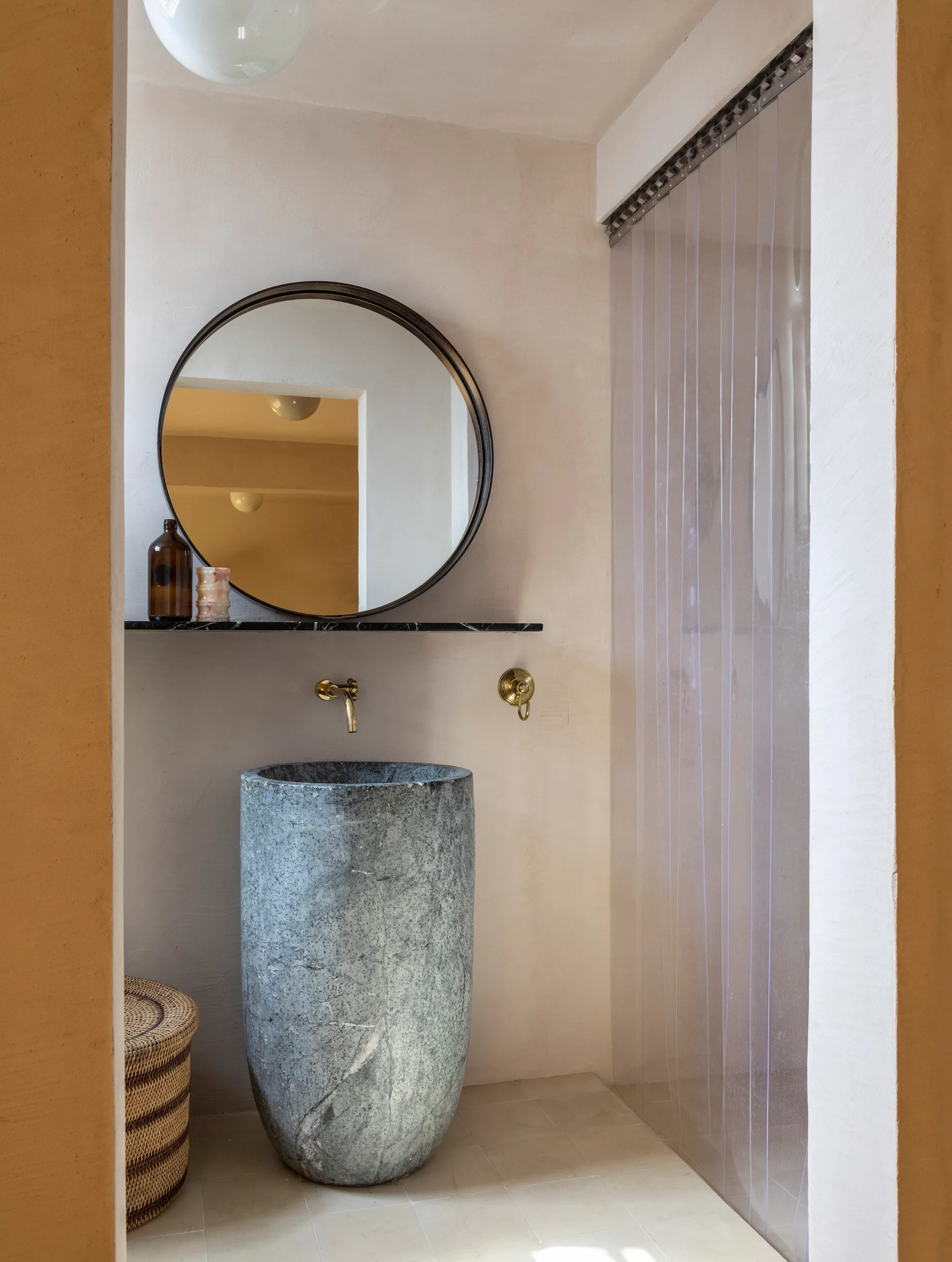 A minimalist bathroom features a textured grey stone pedestal sink, a circular wall mirror, a black stone shelf, and a wall-mounted brass faucet.