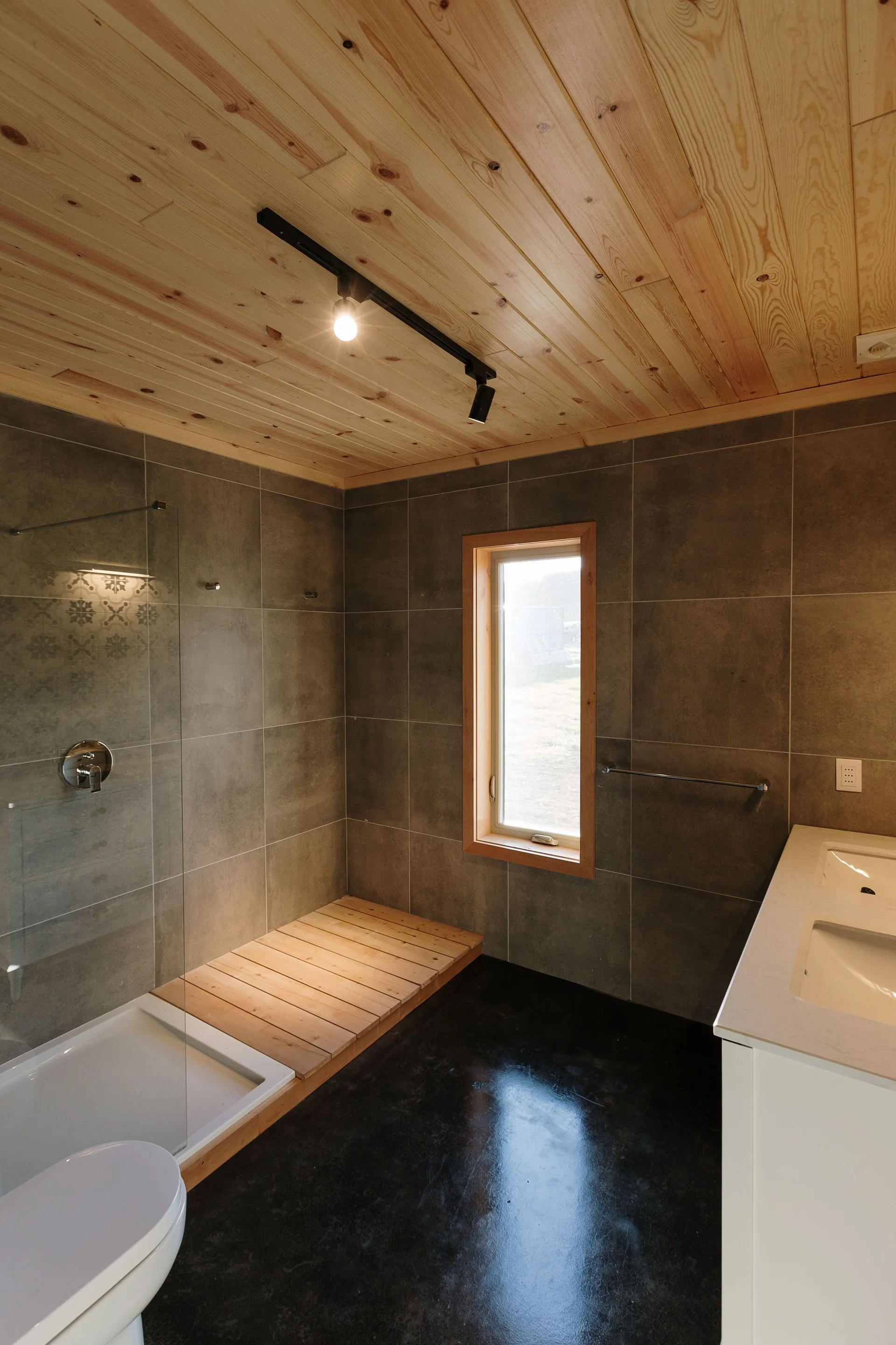 A modern bathroom features a knotty pine plank ceiling, large-format grey tile walls, a white vanity, and a wood-slat shower deck.