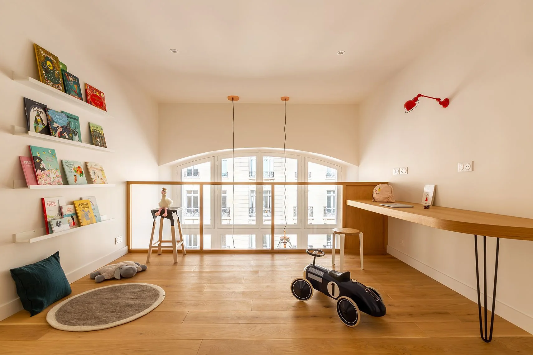A minimalist playroom features light wood floors, a long built-in wooden desk, floating bookshelves on a white wall, and a large arched window.