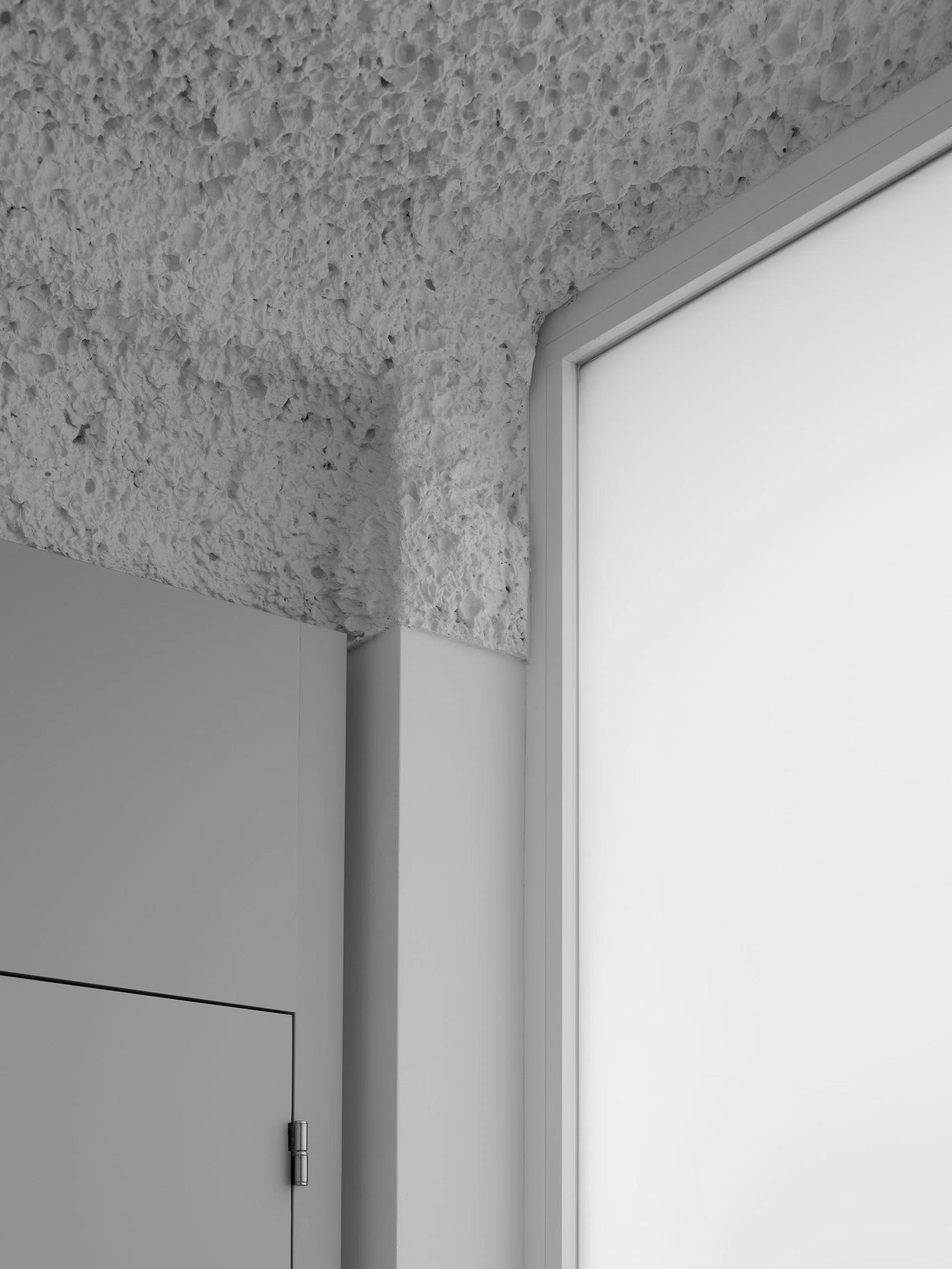 A close-up shows a highly textured gray plaster ceiling meeting a smooth matte gray wall and a minimalist cabinet corner next to a window frame.