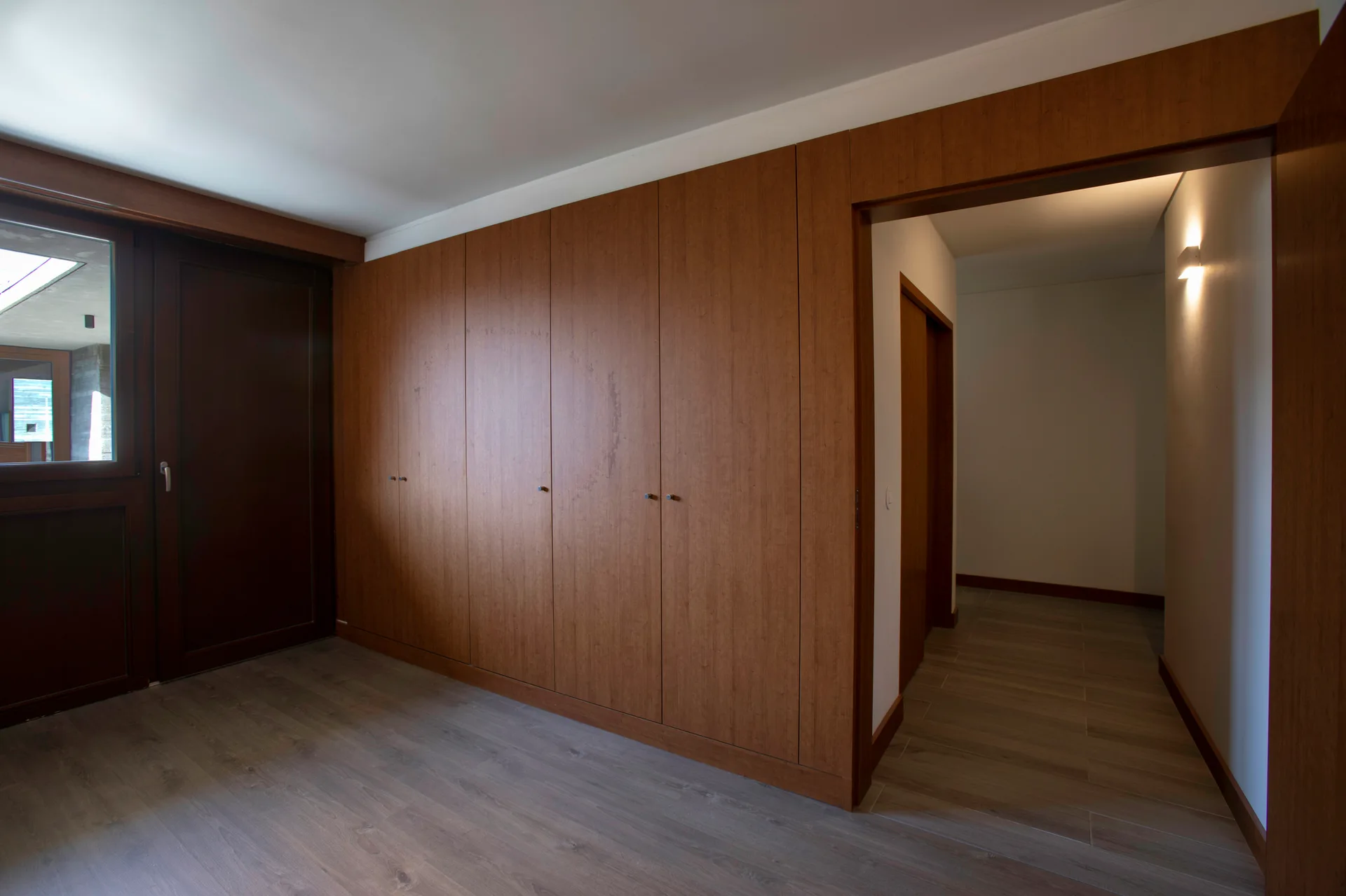A minimalist room features floor-to-ceiling wooden wardrobe cabinets next to an open doorway leading into a lit hallway.