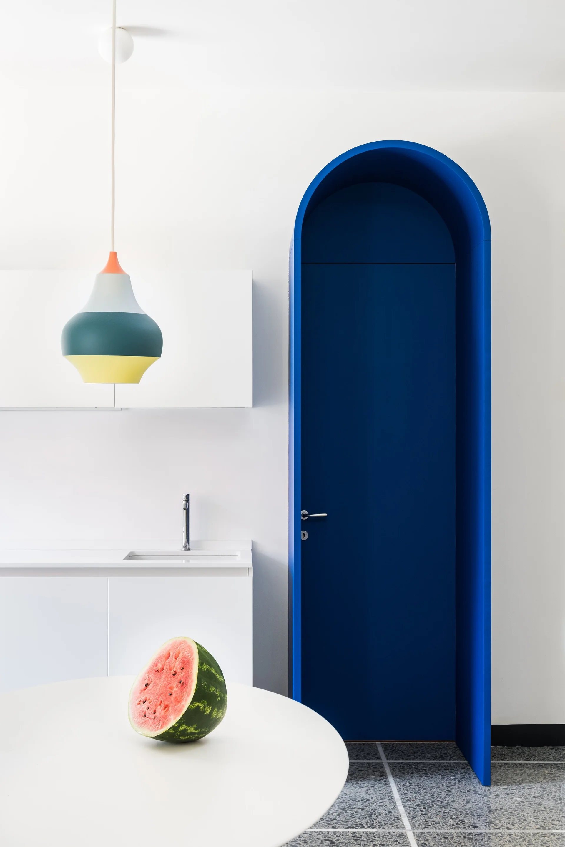 A minimalist white kitchen features a royal blue arched doorway next to plain white cabinets, with a modern pendant light hanging over a foreground table.