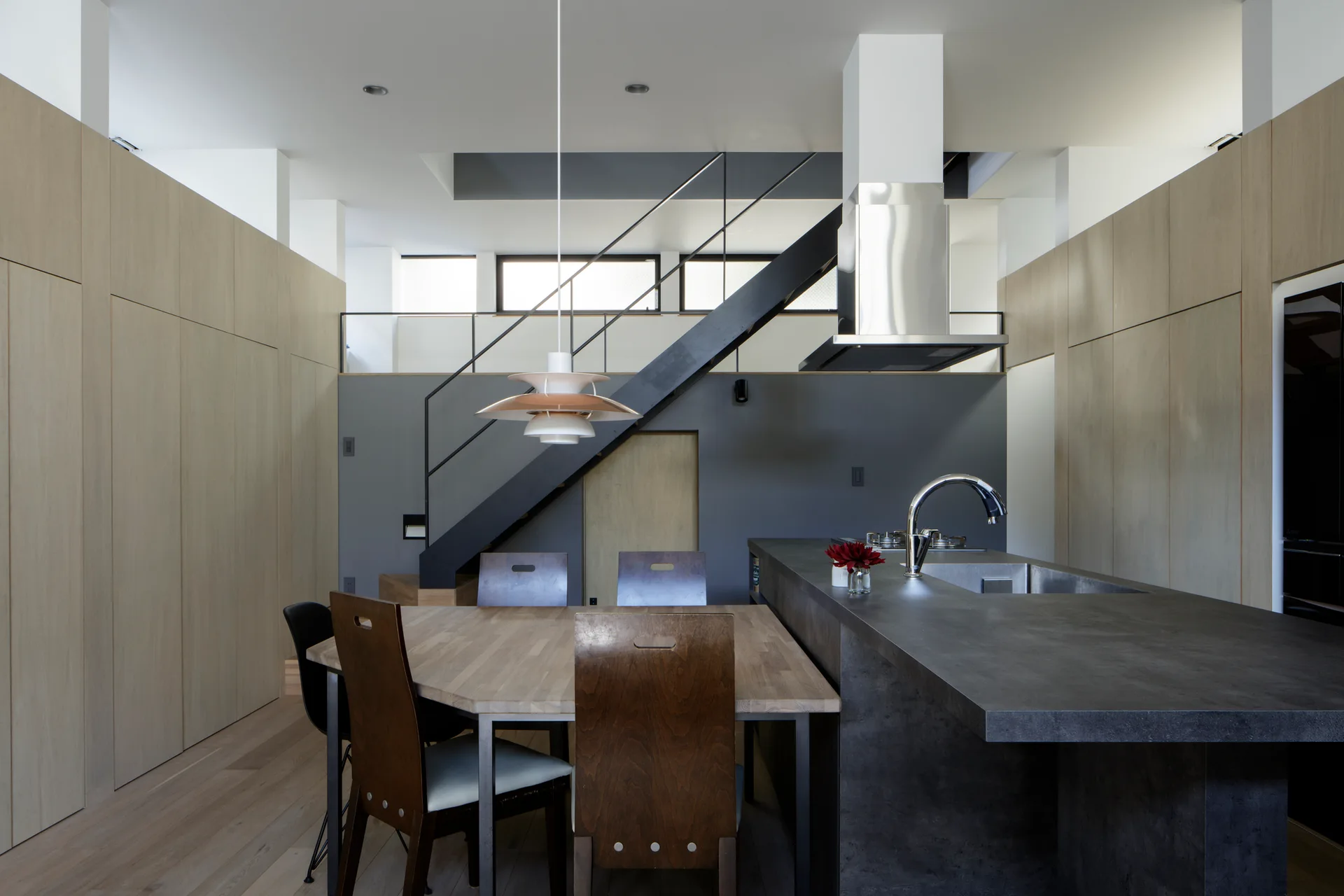 A modern minimalist kitchen features a dark grey island, integrated wood dining table, seamless wood wall panels, and a black steel staircase.