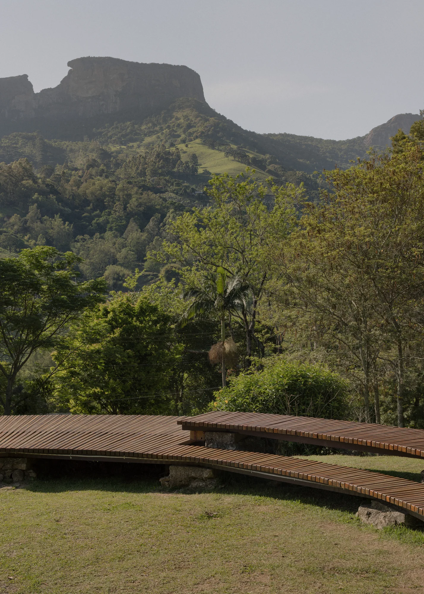 A curved, tiered wooden slat deck sits on a grassy lawn supported by stone bases, with lush trees and a rocky mountain in the background.
