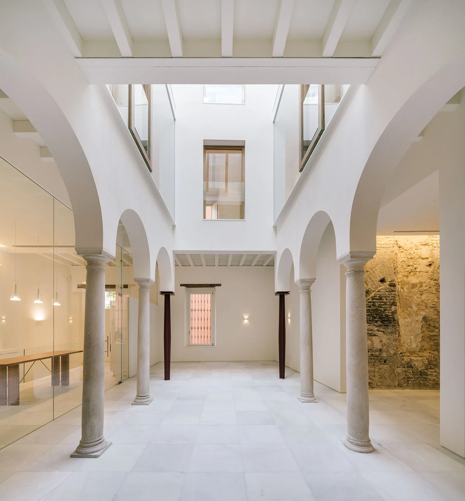 A minimalist white atrium featuring an arched colonnade with stone columns, white flooring, and an exposed historic stone wall.