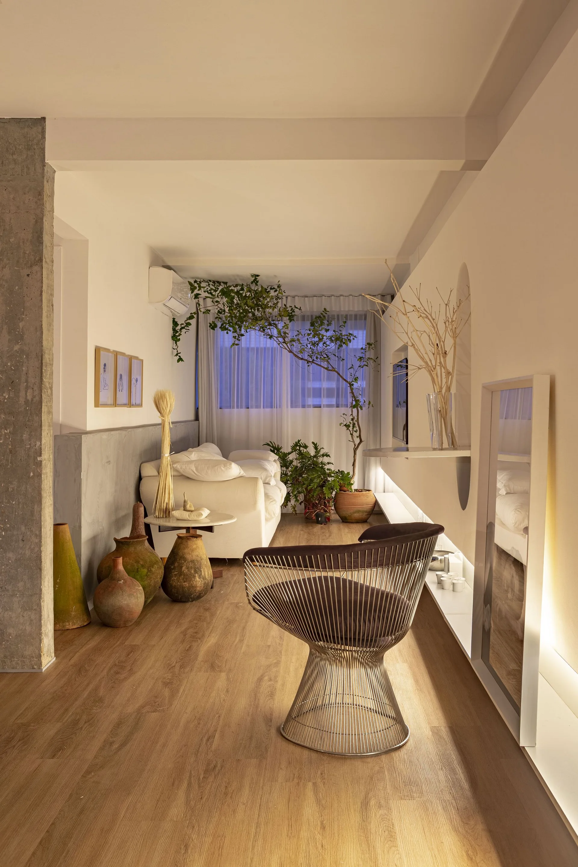A minimalist living room features a metallic wire chair, rustic clay pots, an exposed concrete column, and a low white sofa in the background.