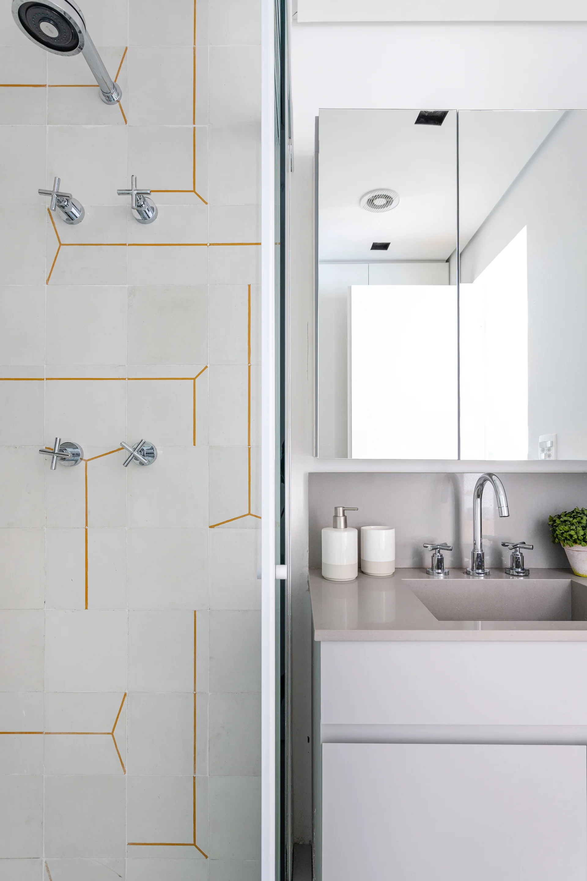 A modern bathroom features a white tiled shower with custom yellow geometric grout lines alongside a light grey vanity.