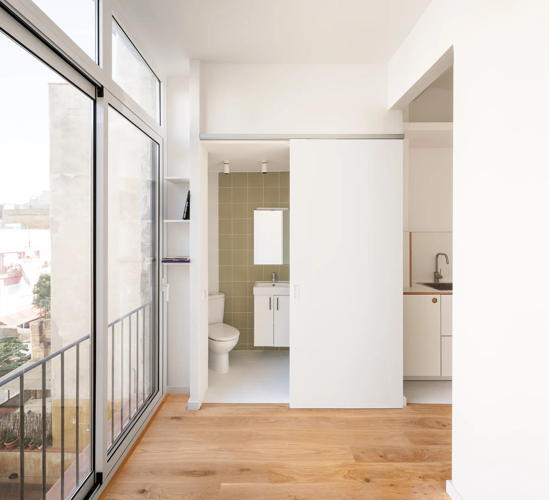 A minimalist studio features a white sliding door opening to a bathroom with olive-green wall tiles, next to light wood floors and large windows.