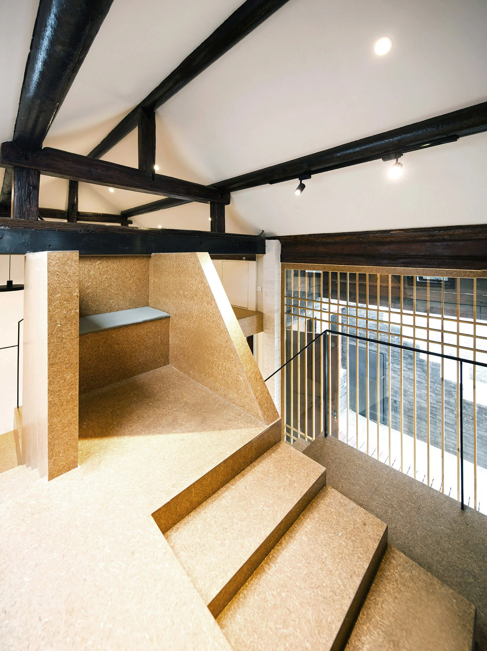 A modern staircase constructed from tan engineered wood panels sits beneath a white vaulted ceiling with dark exposed timber beams.