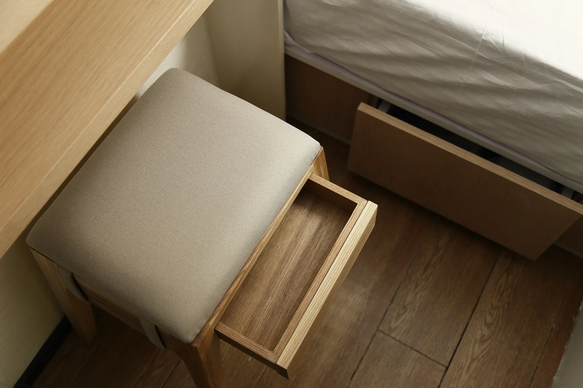 A high-angle view of a light beige upholstered vanity stool with an open wooden storage drawer on a wood floor next to a bed.