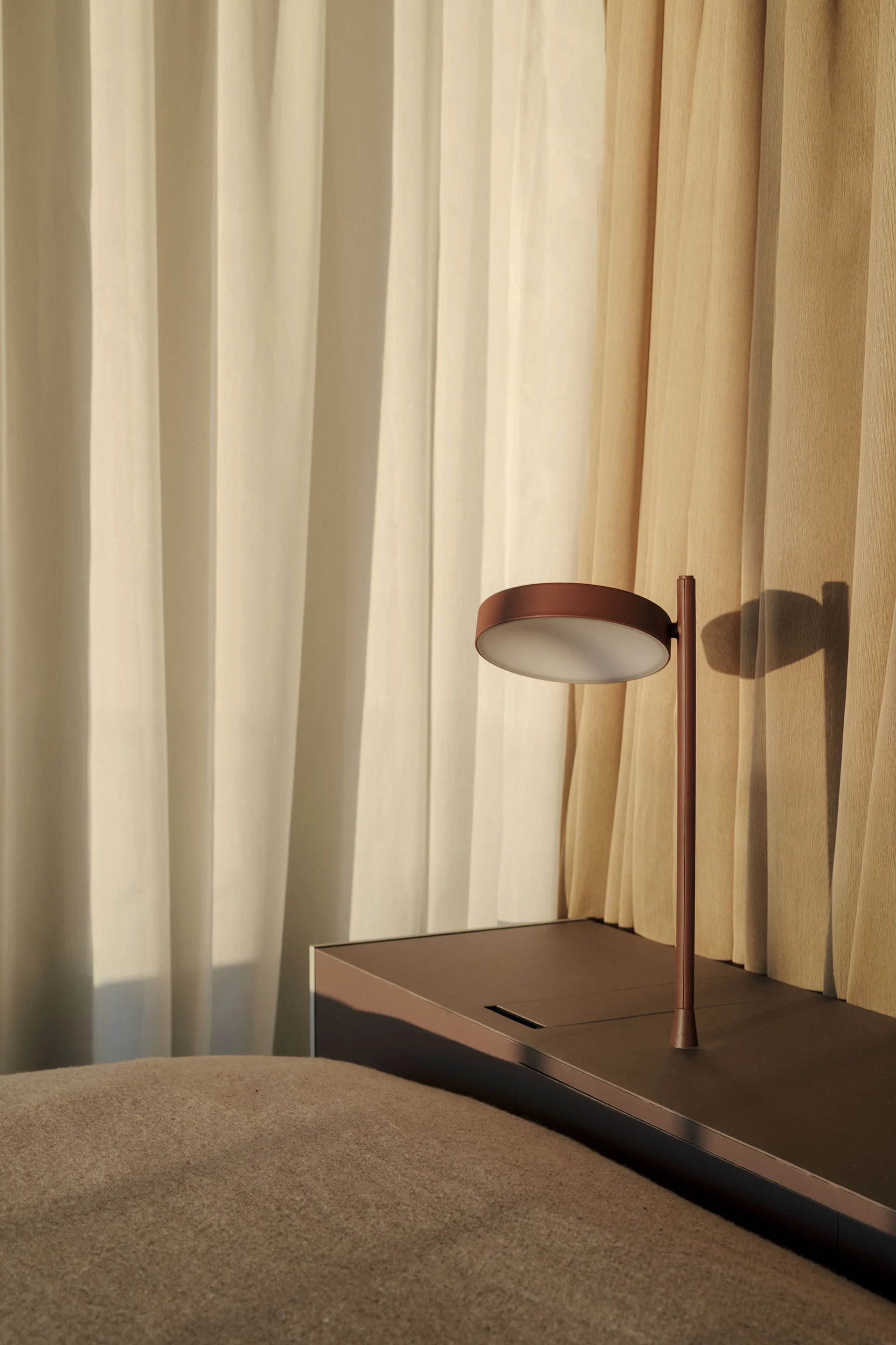 A slim, matte terracotta table lamp with a disc head sits on a dark brown surface in front of layered beige and white pleated curtains.