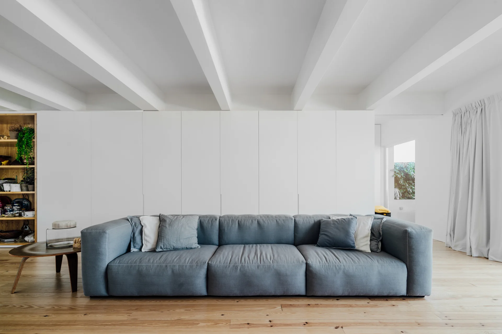 A minimalist living room features a large gray sofa on light wood flooring beneath prominent white ceiling beams and a white storage wall.