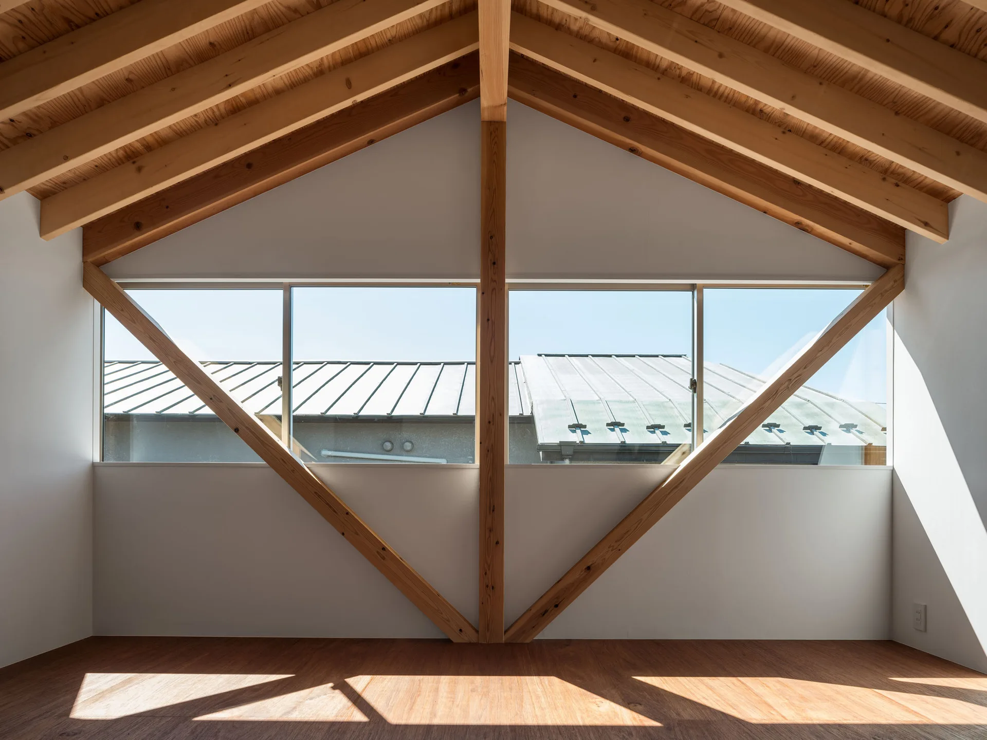 A minimalist attic room with an exposed light wood rafter ceiling, white walls, and a large horizontal window framed by a wooden truss structure.