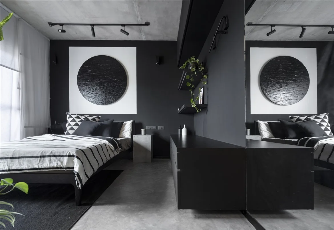 A minimalist monochrome bedroom featuring matte black walls, a raw concrete ceiling, a low bed with striped bedding, and textured circular art.